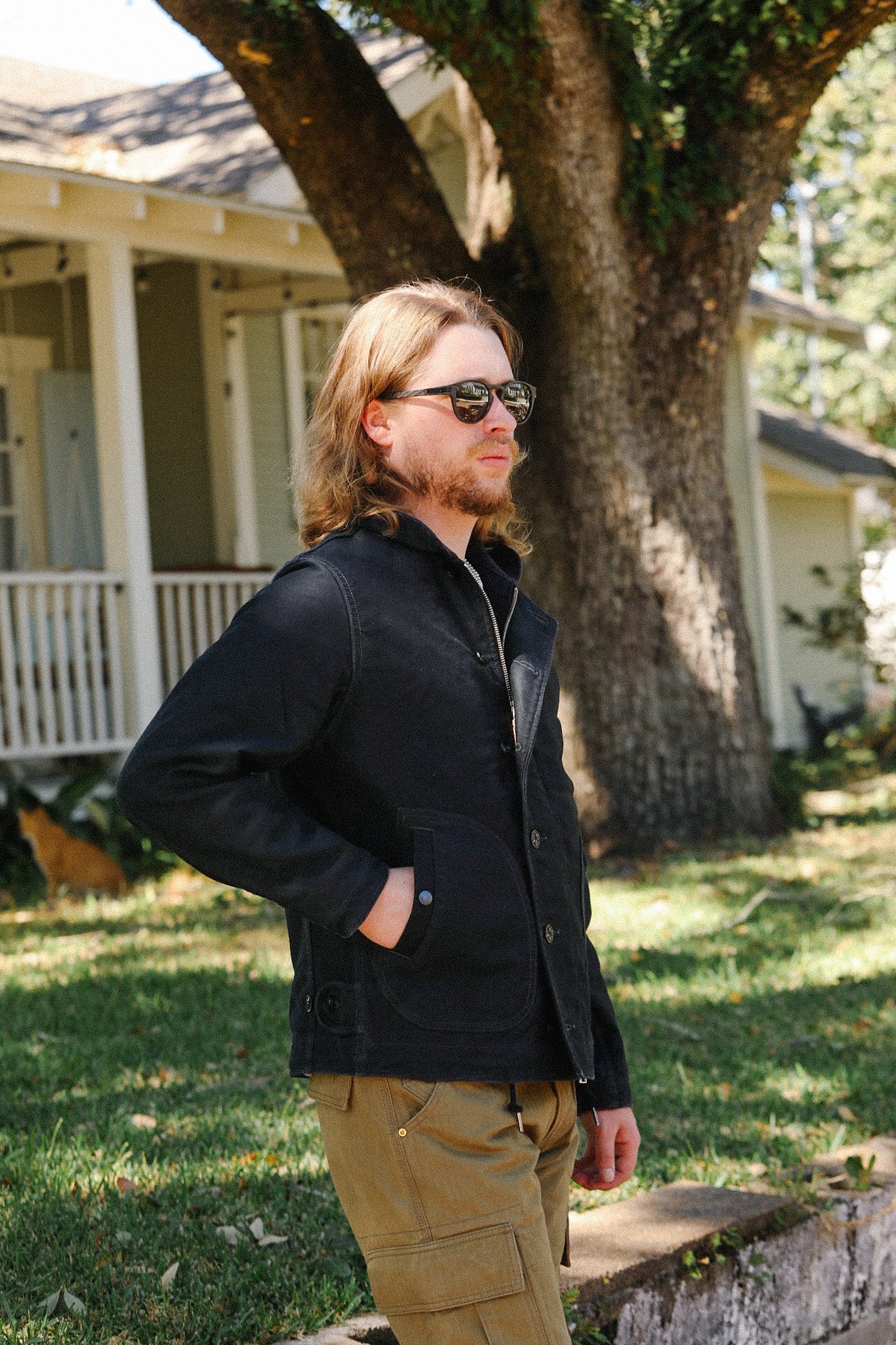Man wearing black jacket and khaki cargo pants standing outdoors near a large tree and house