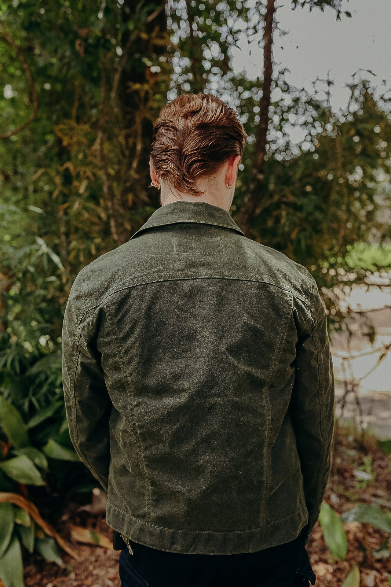 Man wearing green waxed canvas jacket standing outdoors with green foliage background
