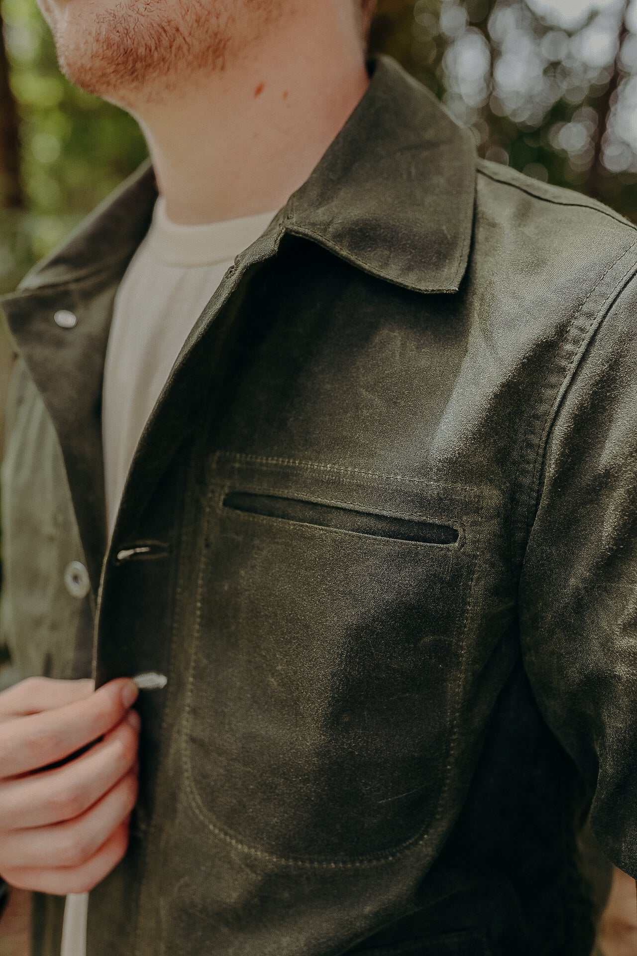 Close-up of a man wearing a dark olive green jacket made of durable fabric, partially buttoned, outdoors