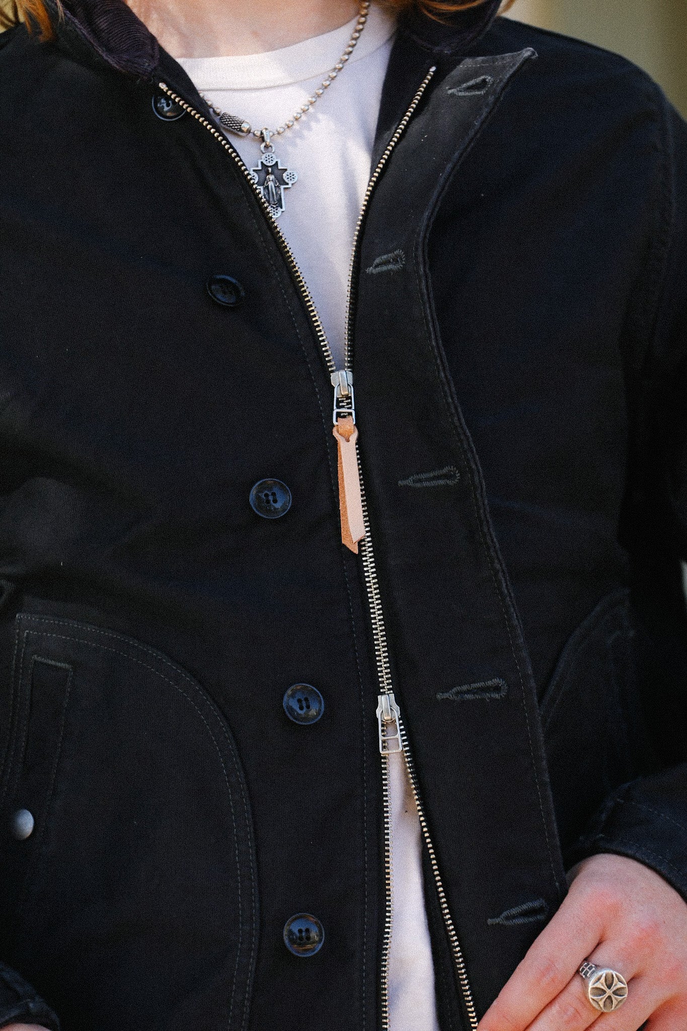 Close-up of person wearing a black zippered jacket over white shirt with silver cross necklace and silver ring