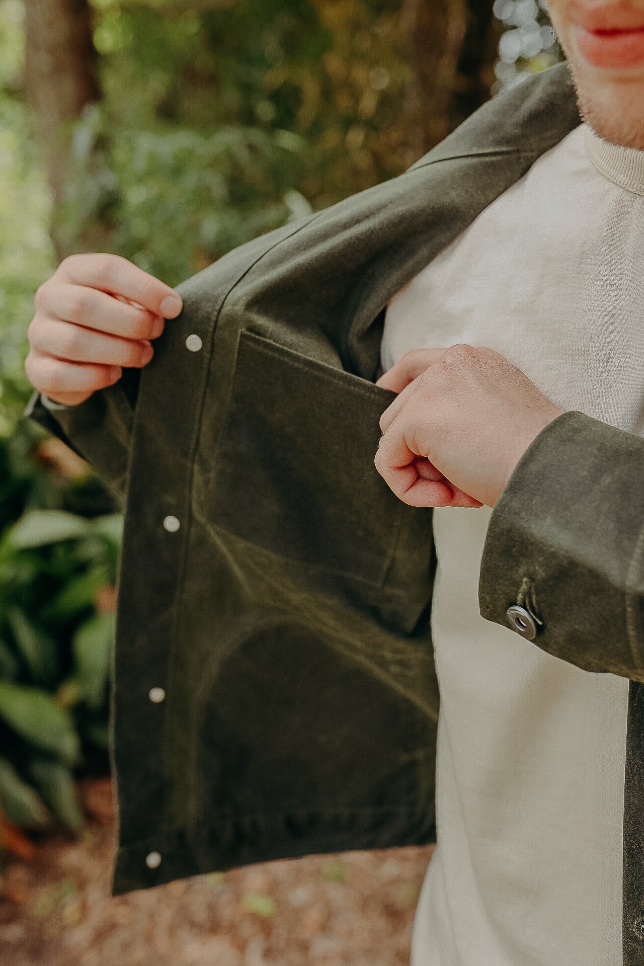 Person opening dark green durable jacket to show inner pocket outdoors with greenery