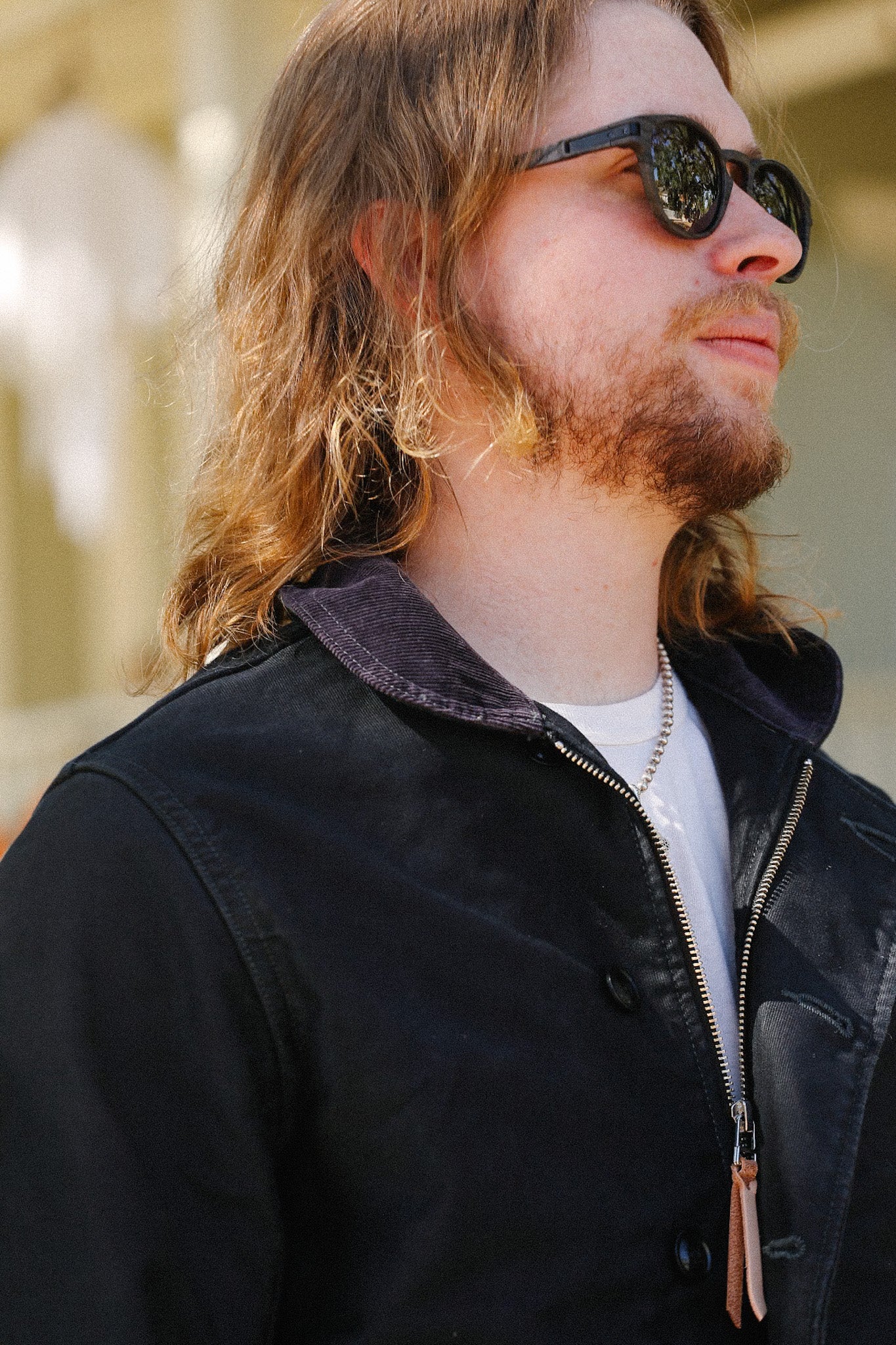 Man with long hair and beard wearing black zip-up jacket, sunglasses, and silver chain necklace