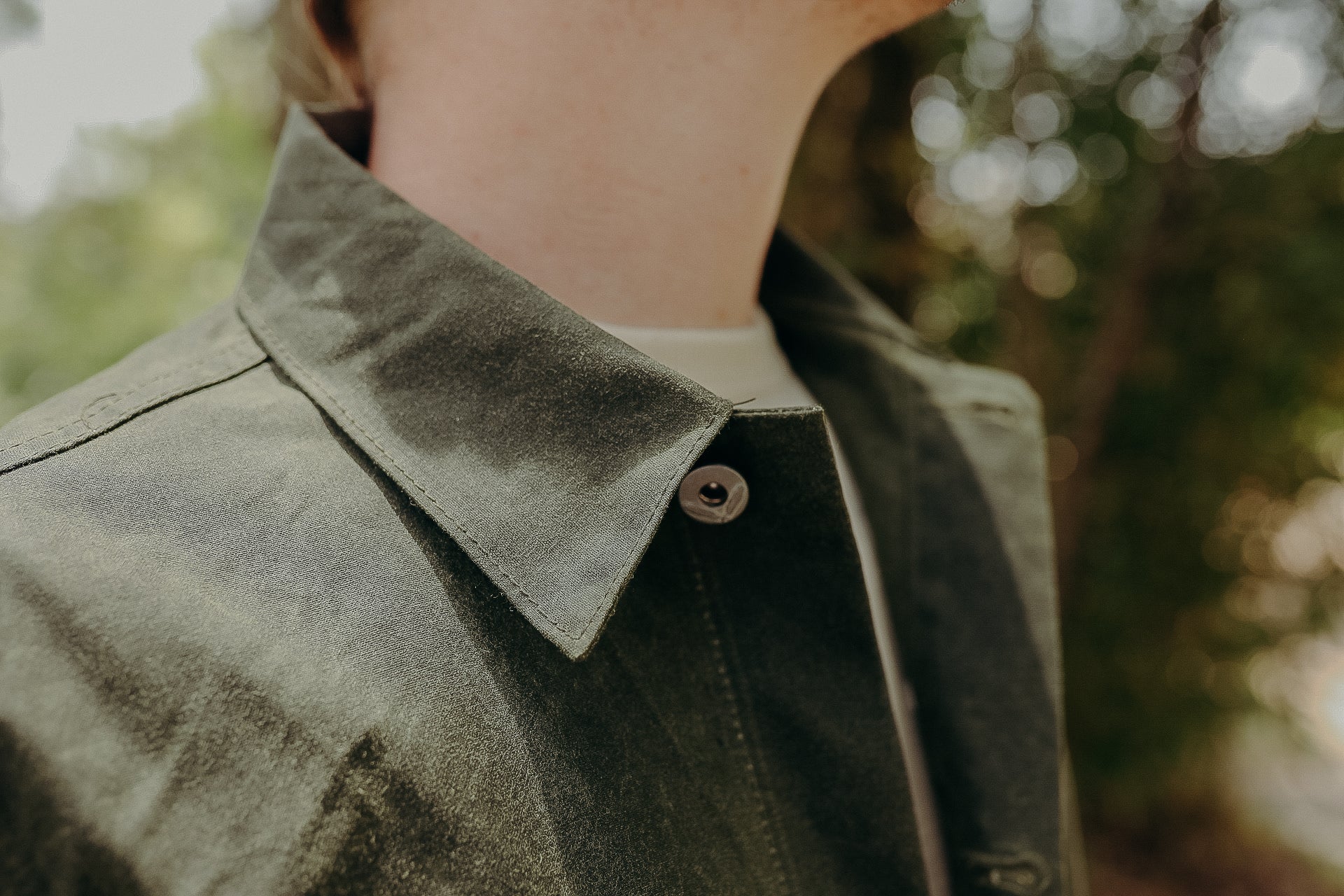 Close-up of olive green button-up shirt collar on person with blurred outdoor foliage background