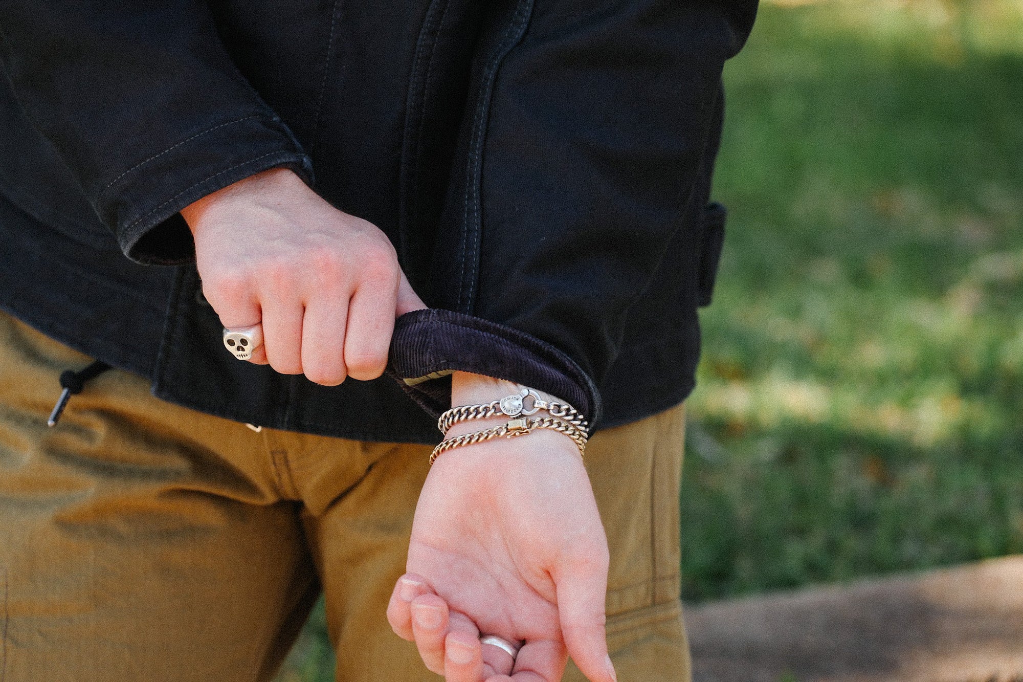 Person wearing silver skull ring and chain bracelets with black jacket and brown pants outdoors