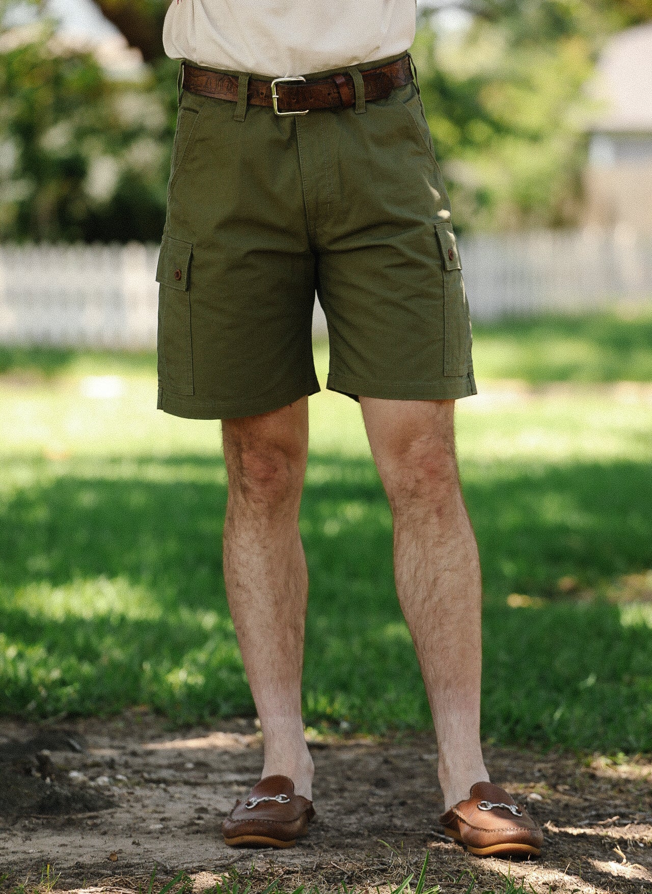 Man wearing olive green cargo shorts with brown leather belt and brown loafers standing outdoors on grass