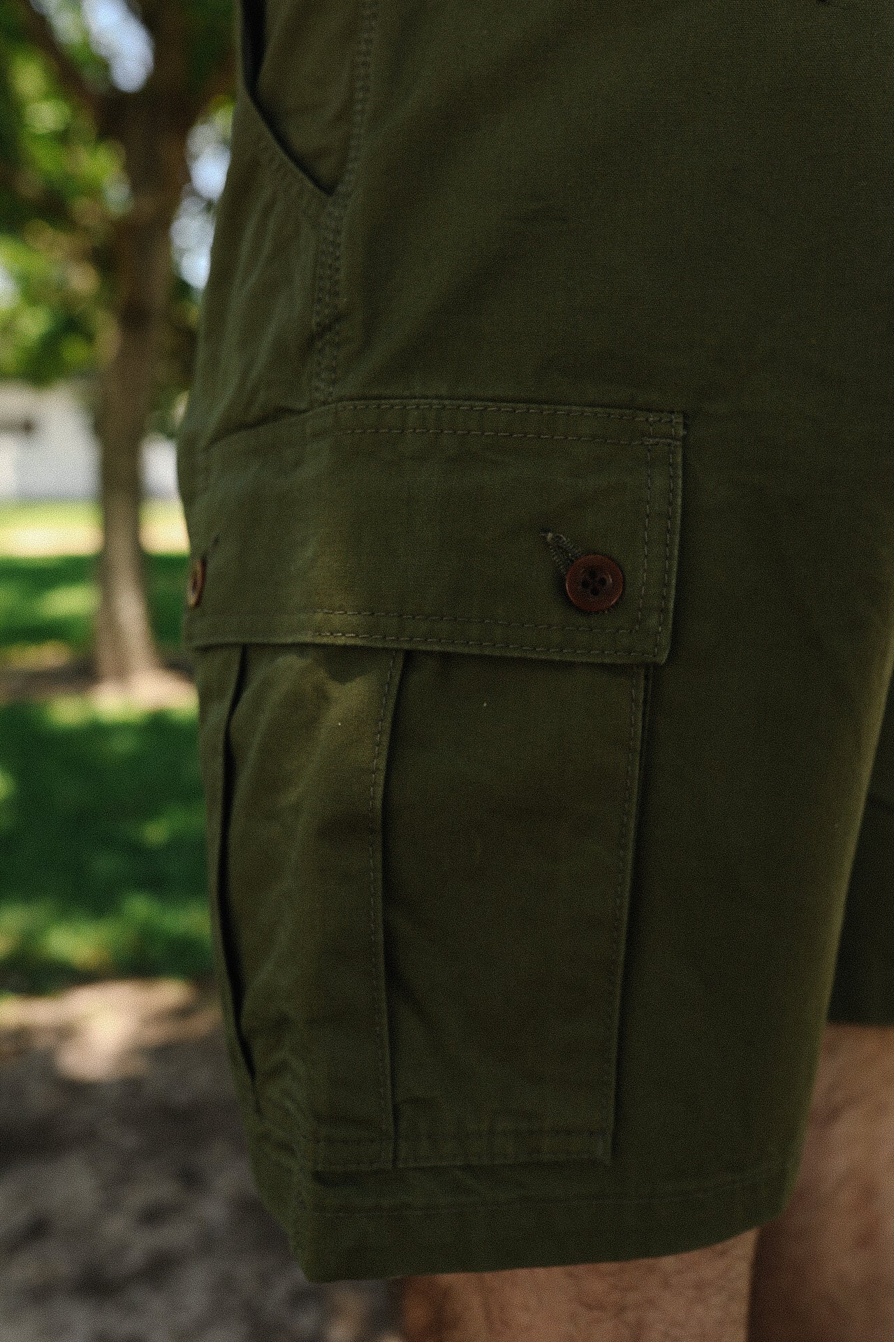 Close-up of olive green cargo shorts pocket with button closure, outdoor setting with grass and trees