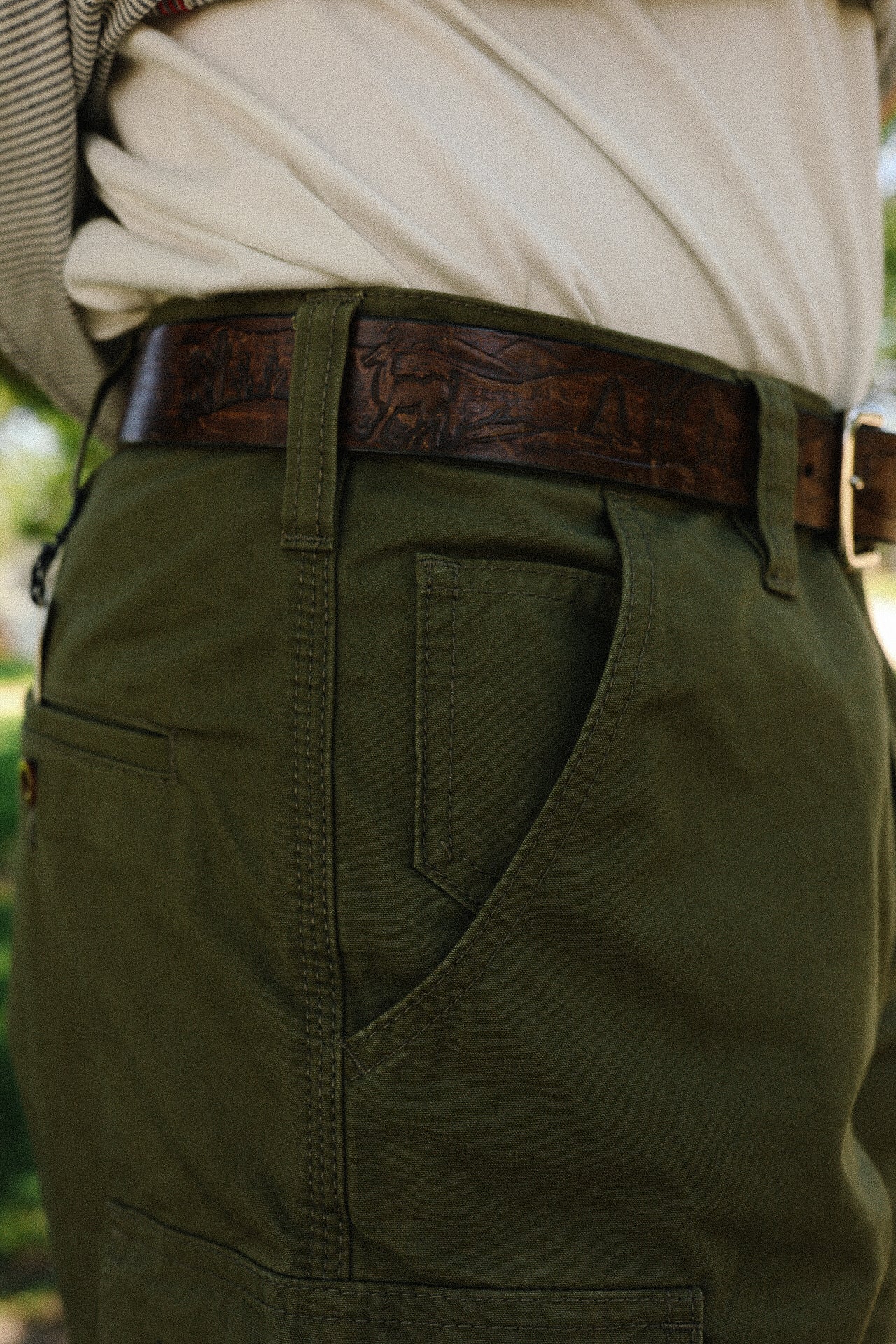 person wearing olive green utility pants and a dark brown embossed leather belt outdoors