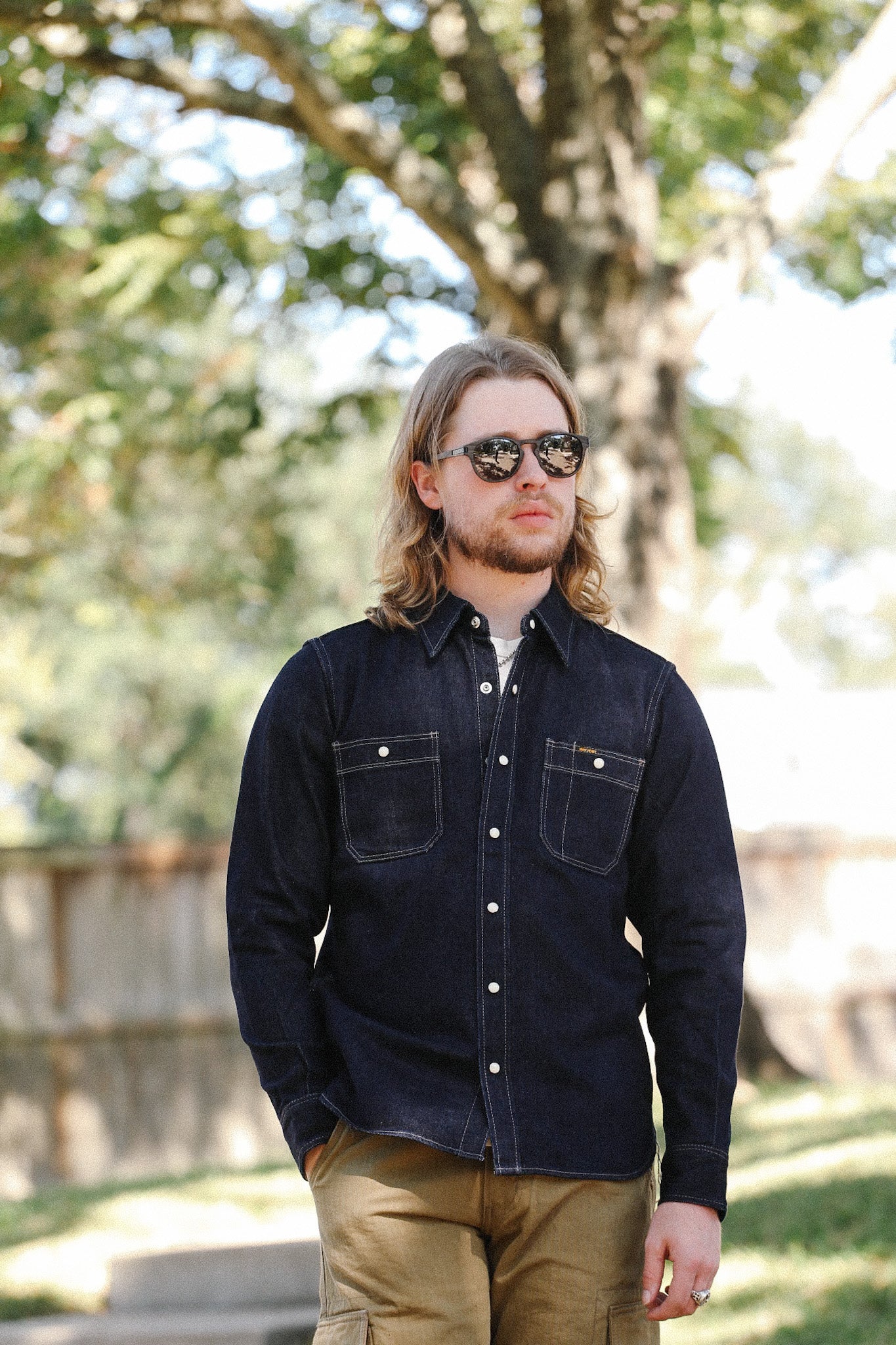 Man wearing dark denim work shirt and sunglasses outdoors with tree and fence background