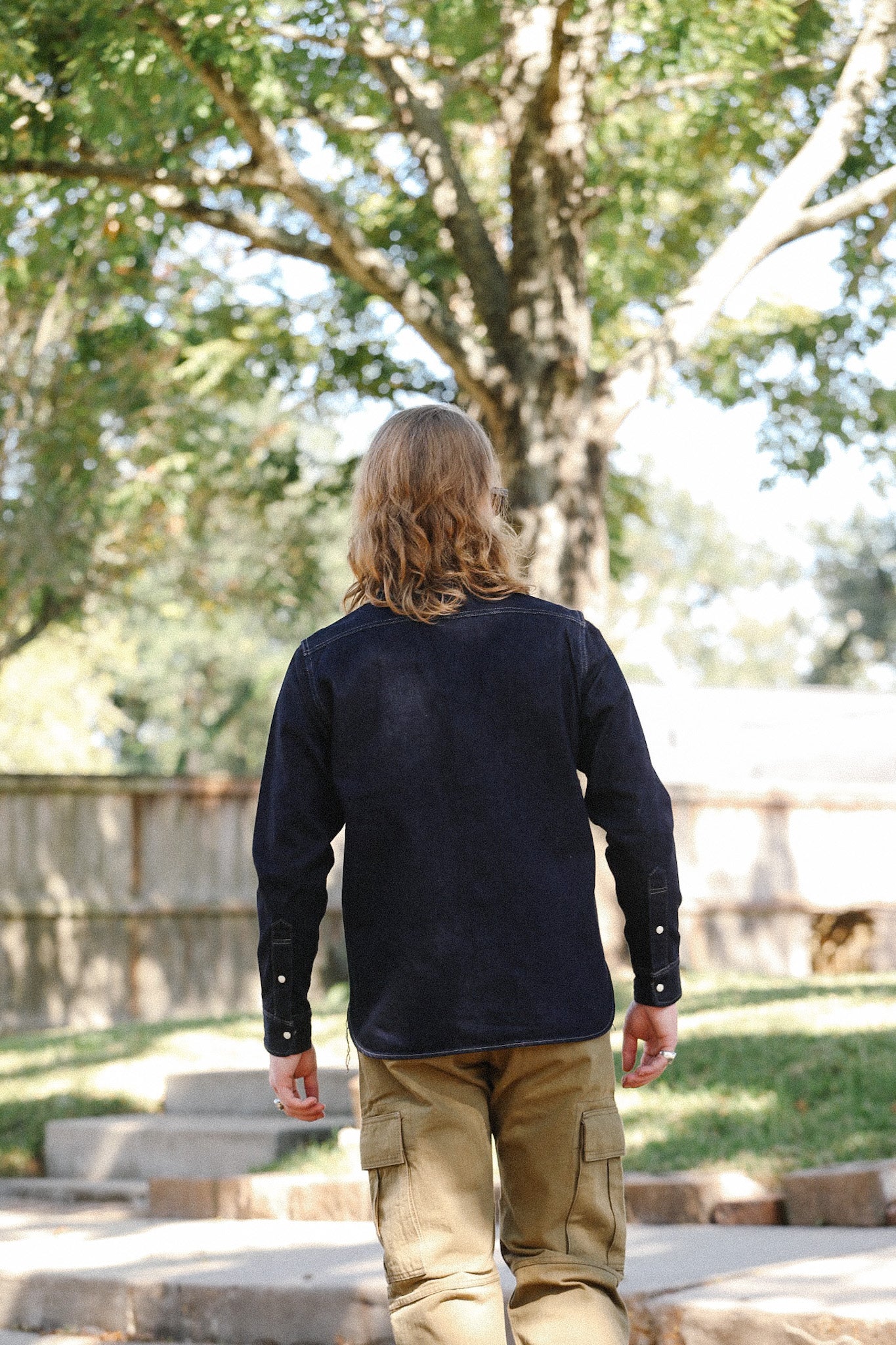 Man with long hair wearing a black jacket and khaki cargo pants walking outdoors in a sunlit garden