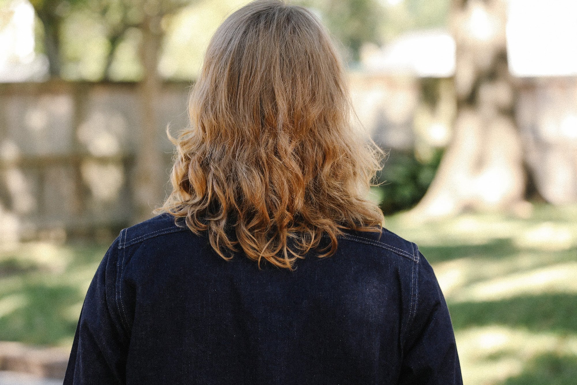 Person with medium-length wavy blonde hair wearing a dark jacket outdoors in a sunny garden