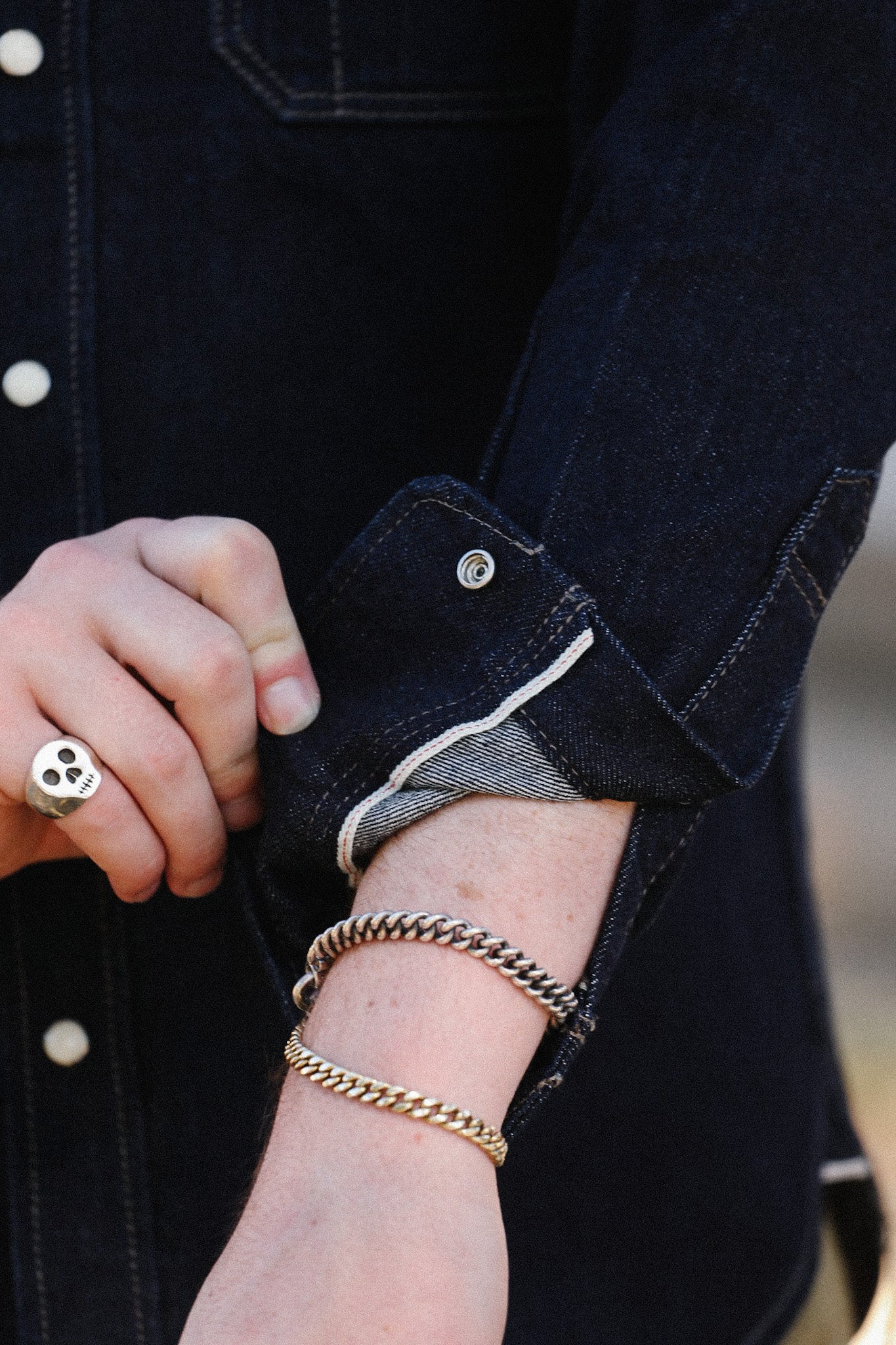 Close-up of hand with skull ring and two chain bracelets wearing dark denim jacket sleeve
