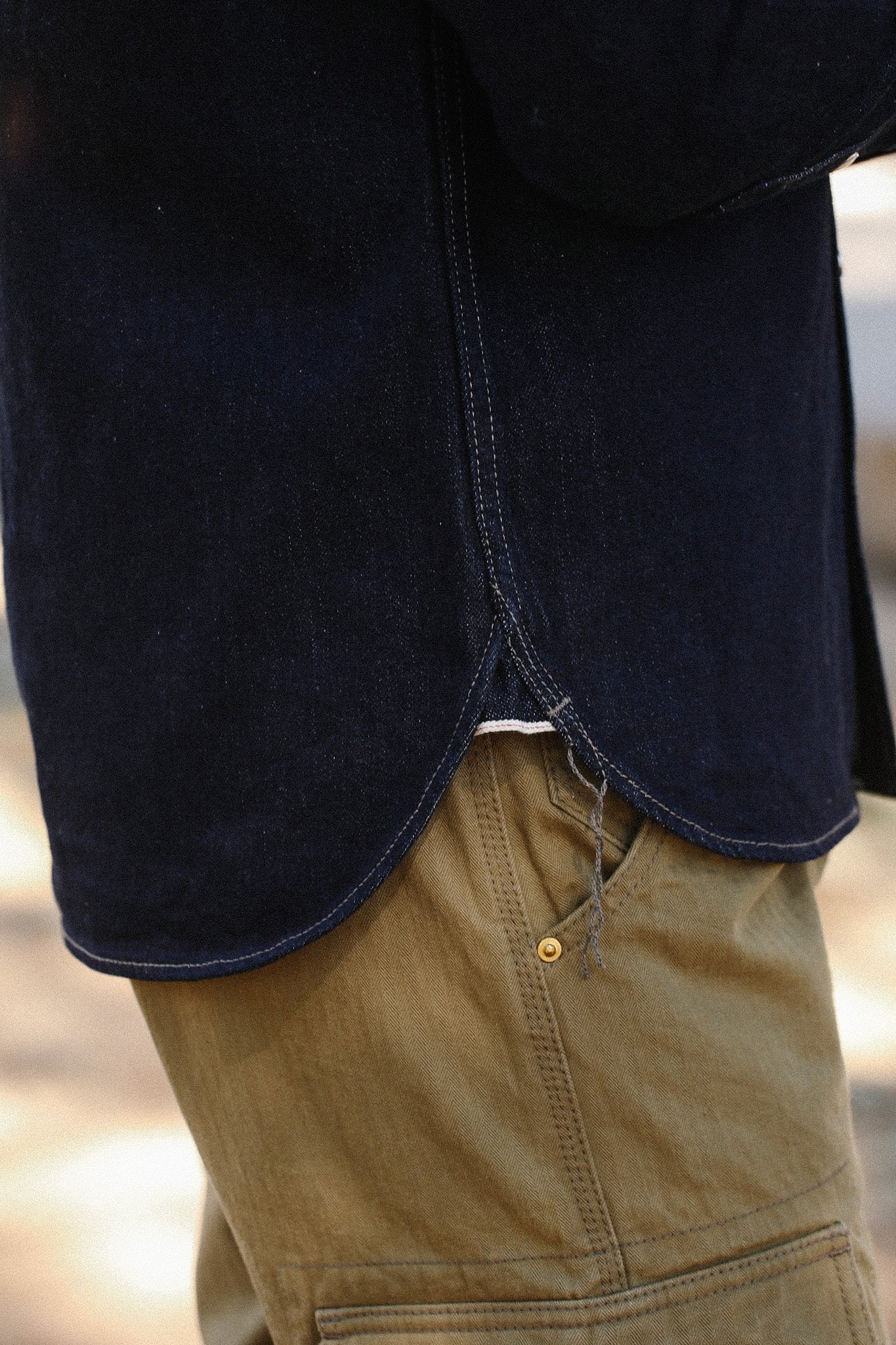 Close-up of navy blue shirt hem with raw edge detail over tan work pants with brass rivet