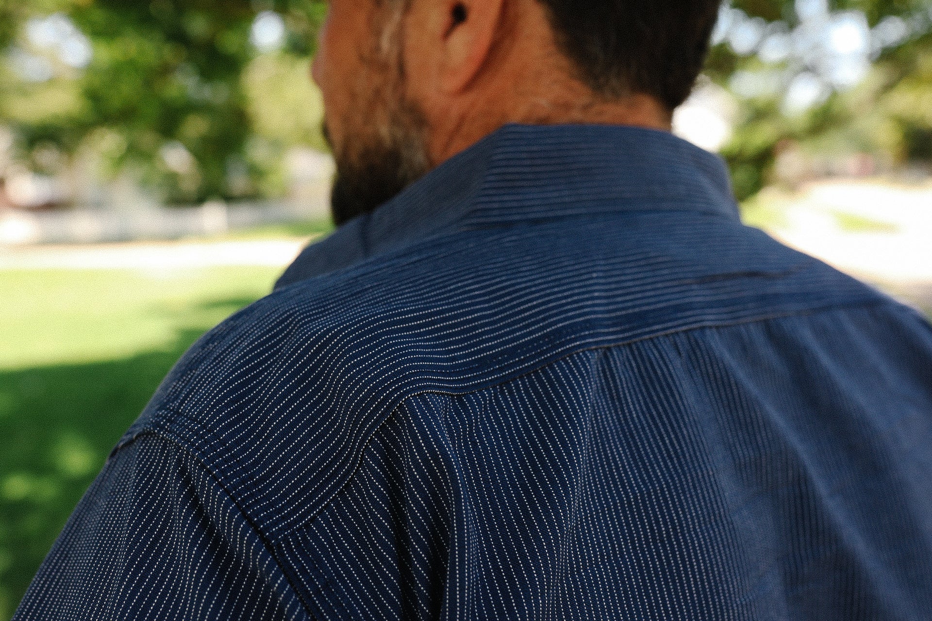 Close-up of a man wearing a navy blue striped work shirt outdoors with blurred green trees in the background