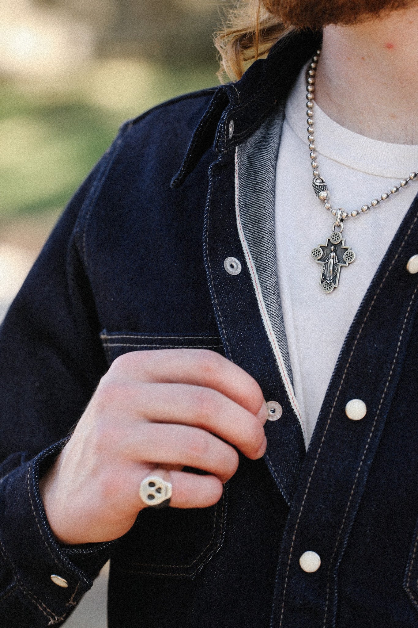 Close-up of person wearing a denim jacket, white t-shirt, silver cross necklace, and skull ring
