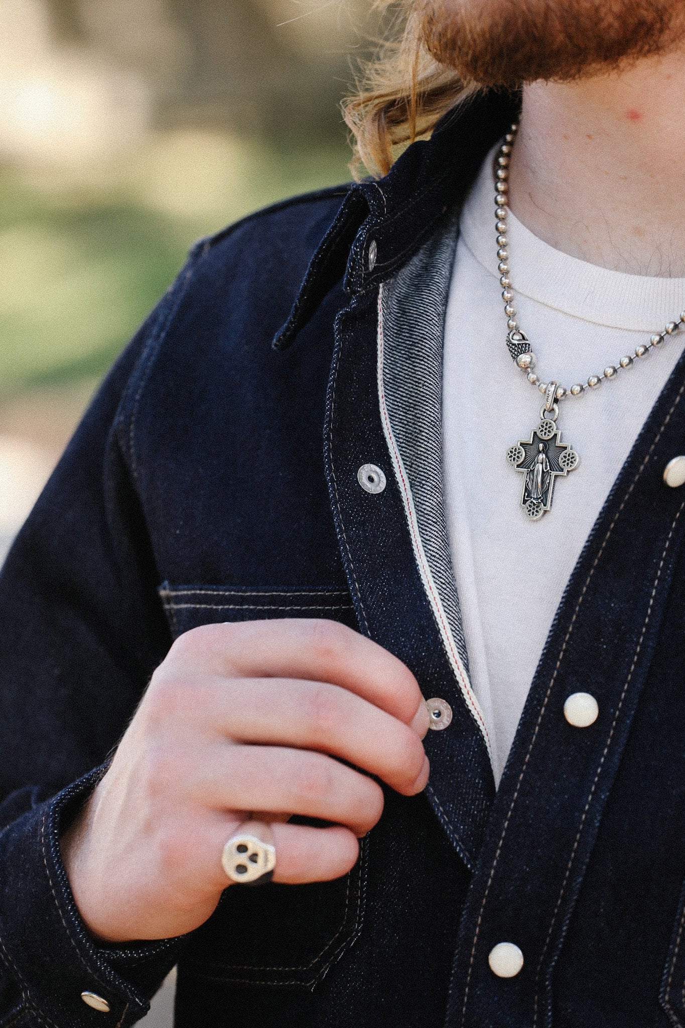 Close-up of person wearing denim jacket, white shirt, silver cross necklace, and skull ring