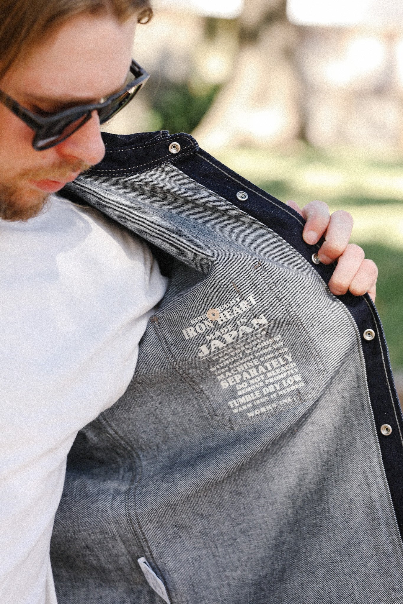 Man wearing sunglasses showing the inside label of a denim jacket by Iron Heart, made in Japan