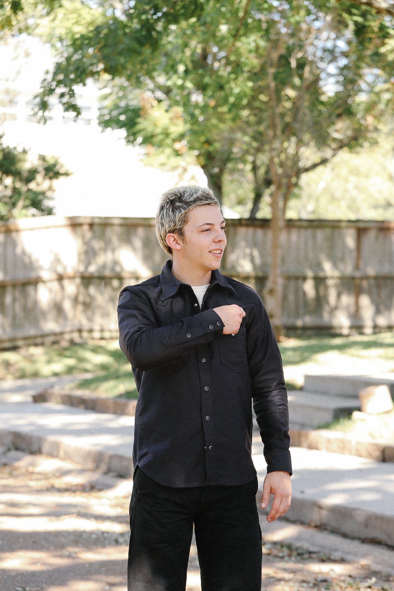 Young man wearing black button-up shirt and pants outdoors near wooden fence and trees
