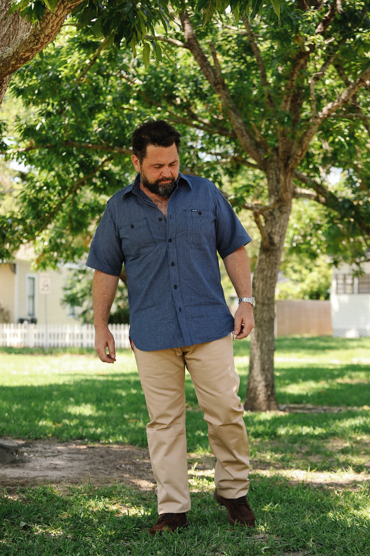 Man wearing blue short-sleeve work shirt and tan pants standing under a tree in a park