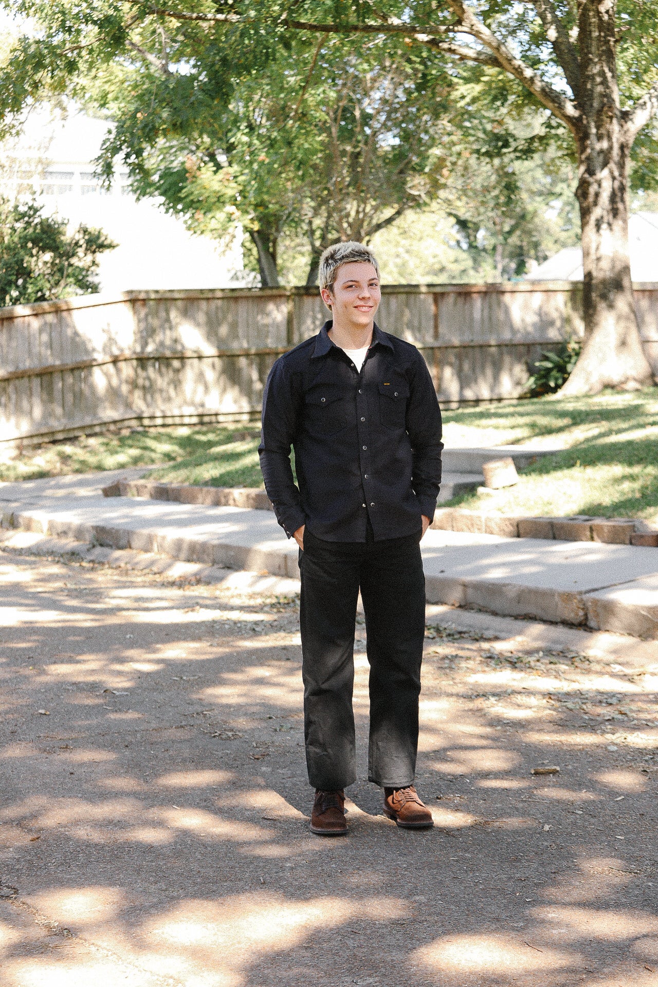 Young person wearing black workwear shirt, black pants, and brown boots standing outdoors on sunlit path