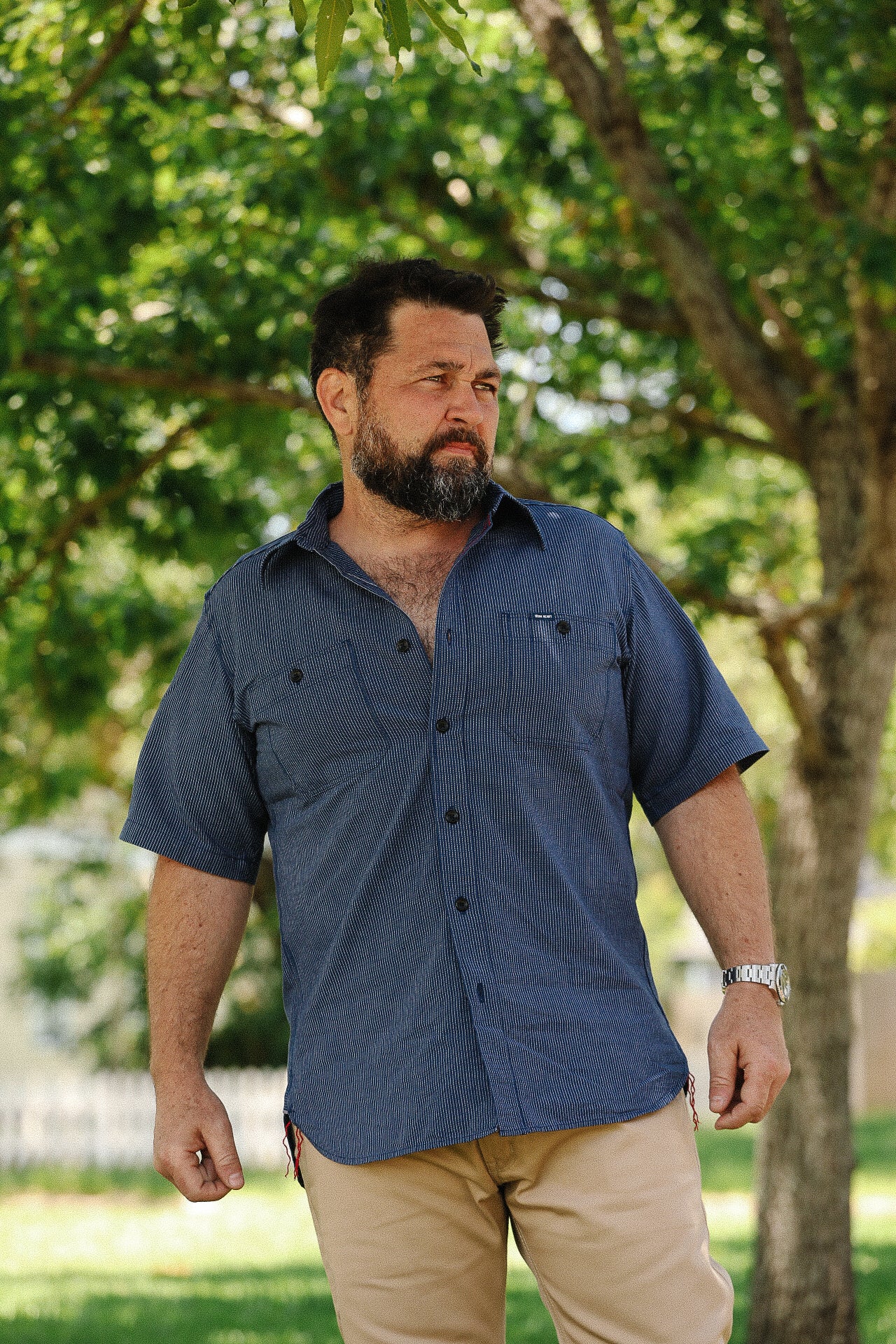 Bearded man wearing a short-sleeve blue button-up shirt and tan pants standing outdoors with green trees