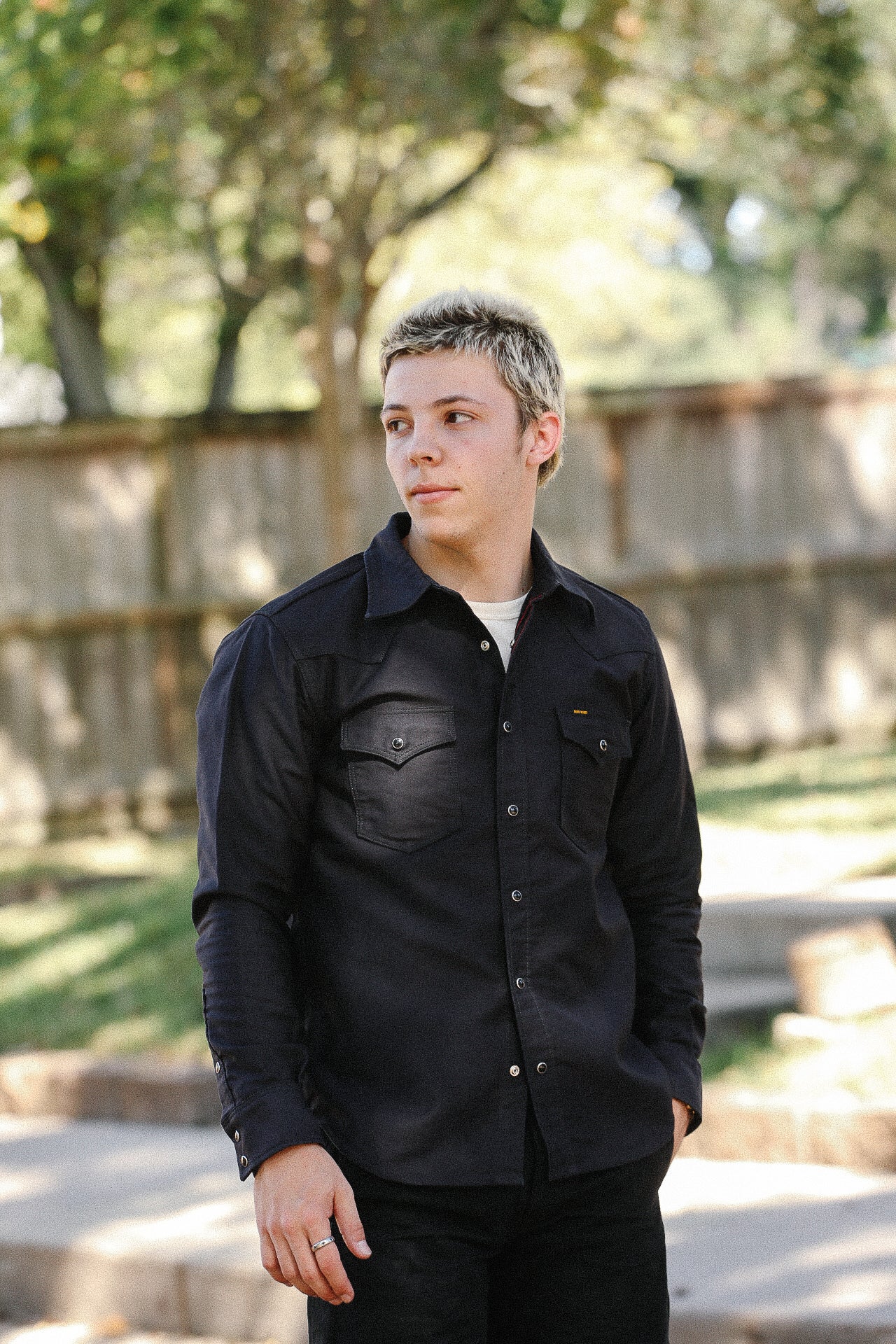 Young man wearing black button-up shirt with chest pockets standing outdoors near wooden fence