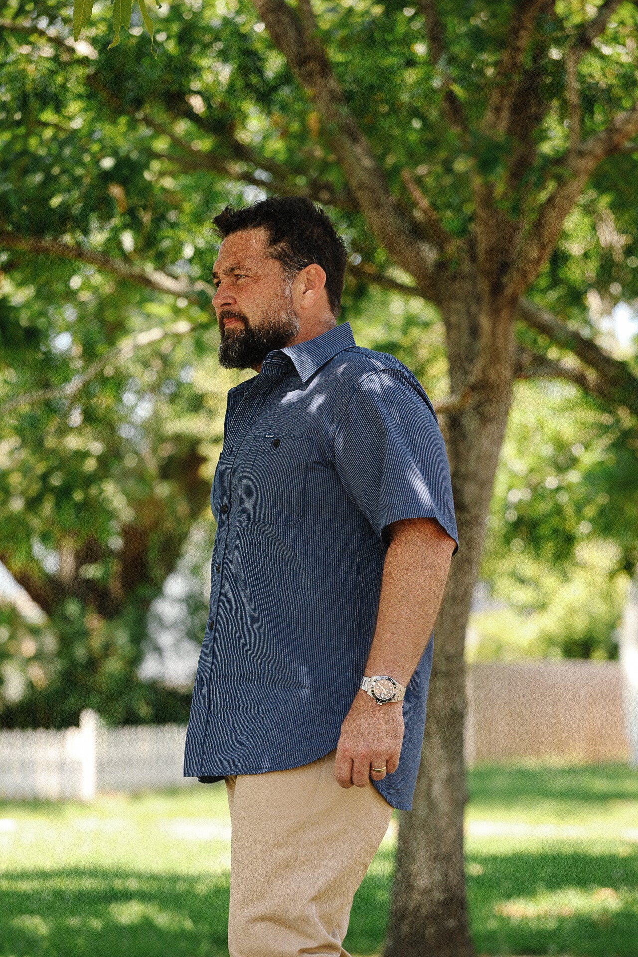 Bearded man wearing a blue short-sleeve button-up shirt and beige pants standing outdoors by a tree
