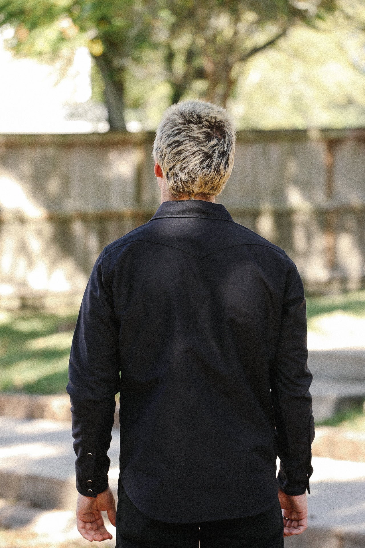 Man with short blond hair wearing a black long-sleeve shirt standing outdoors near wooden fence
