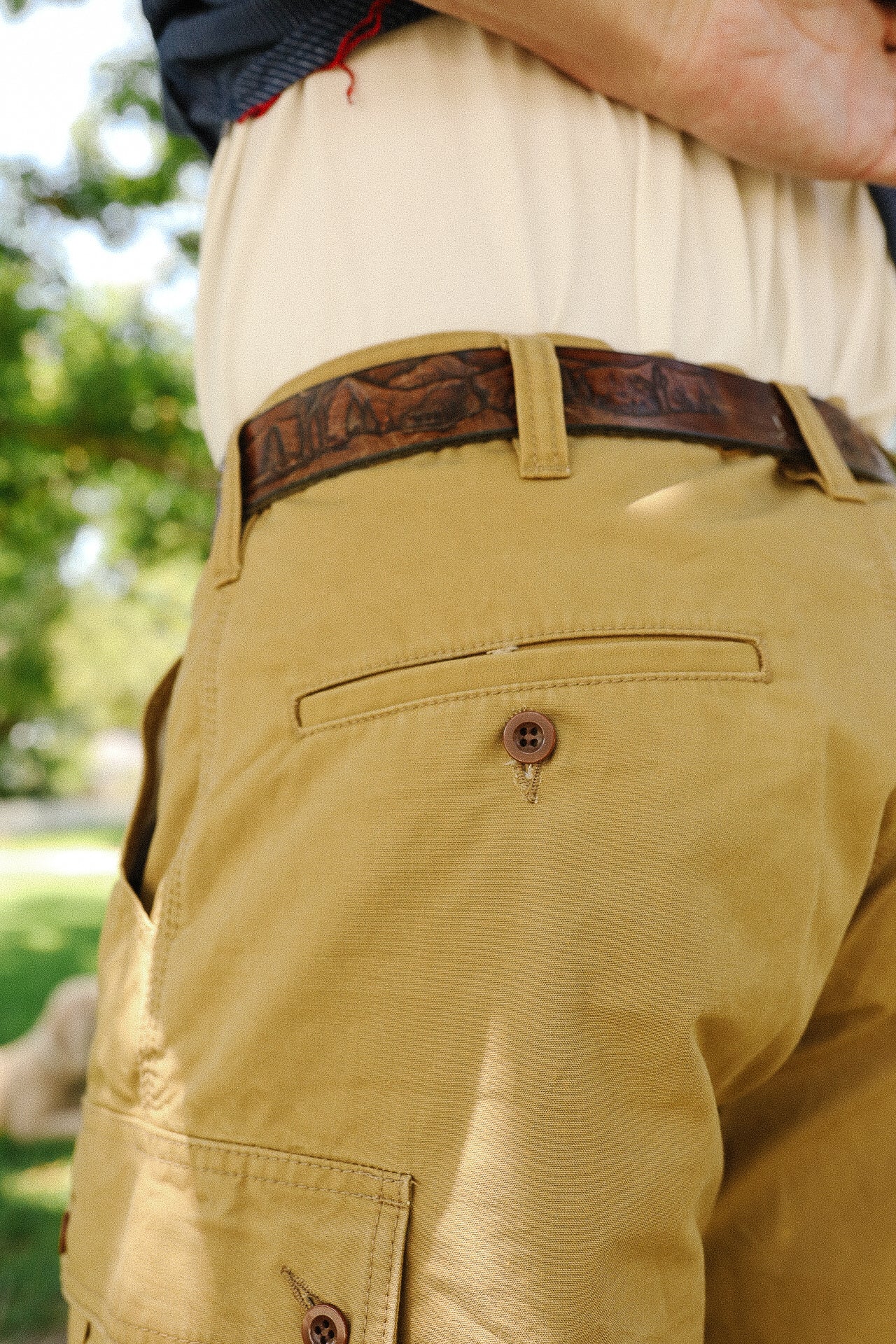 Close-up of a person wearing tan work pants with buttoned back pocket and a brown embossed leather belt outdoors