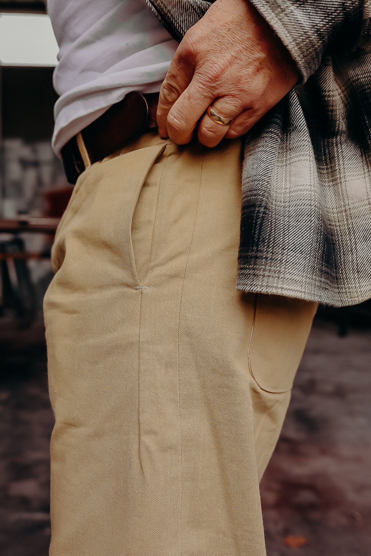 Close-up of a person wearing beige work pants and a plaid shirt, hand with gold ring tucked in pocket