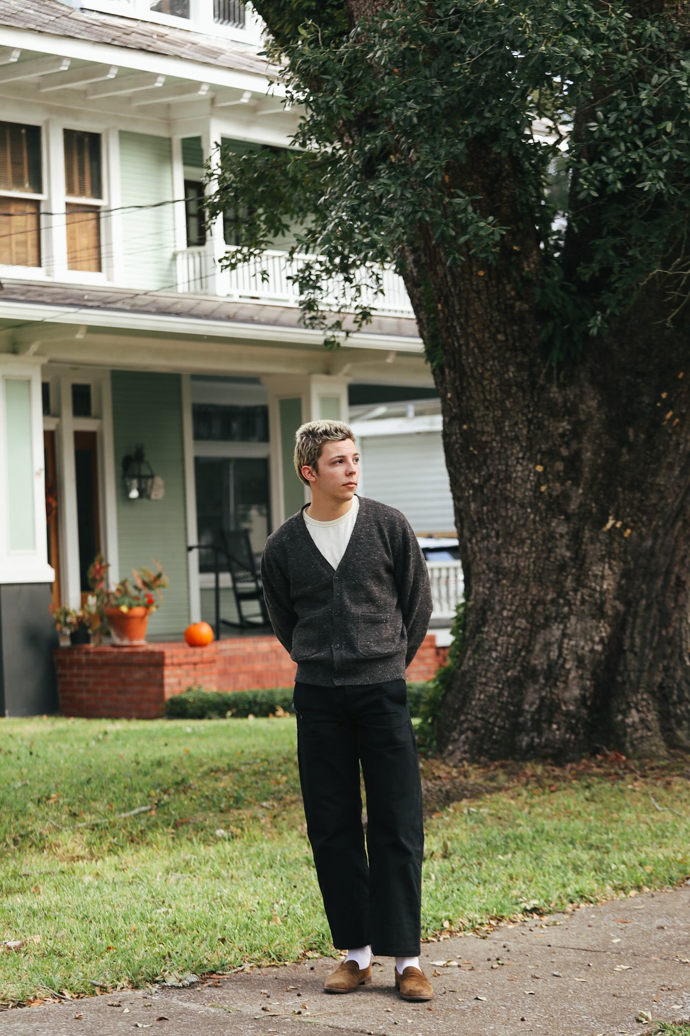 Young man in dark cardigan, white shirt, black pants, and brown shoes standing by large tree in front of green house with pumpkin on porch