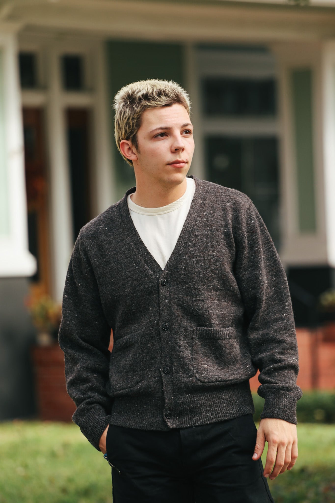 Young man wearing dark buttoned cardigan and white shirt outdoors near house with green lawn
