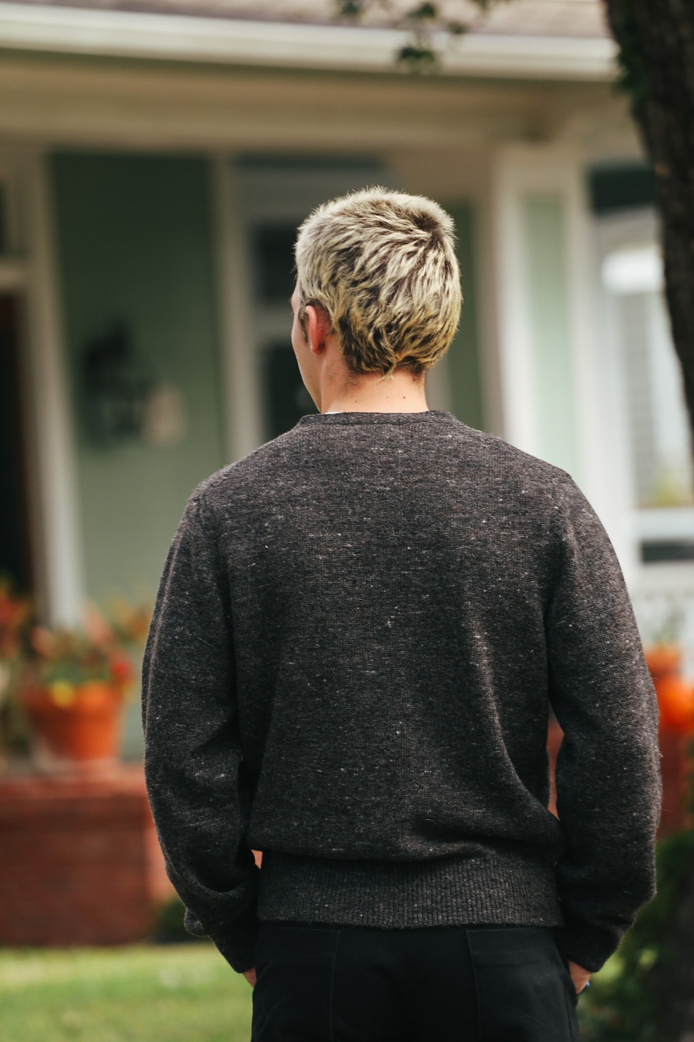Person with short blond hair wearing a dark gray sweater standing outdoors near a house