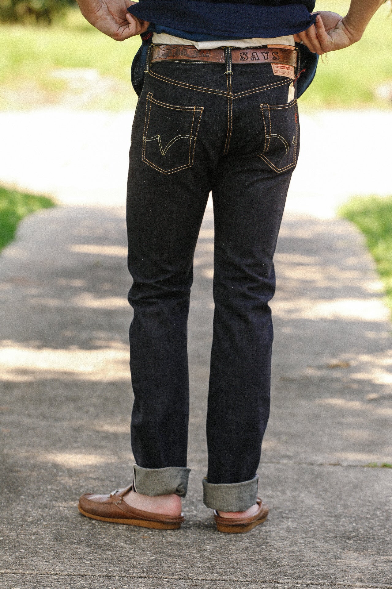 Back view of person wearing dark denim jeans with rolled cuffs and brown leather loafers outdoors