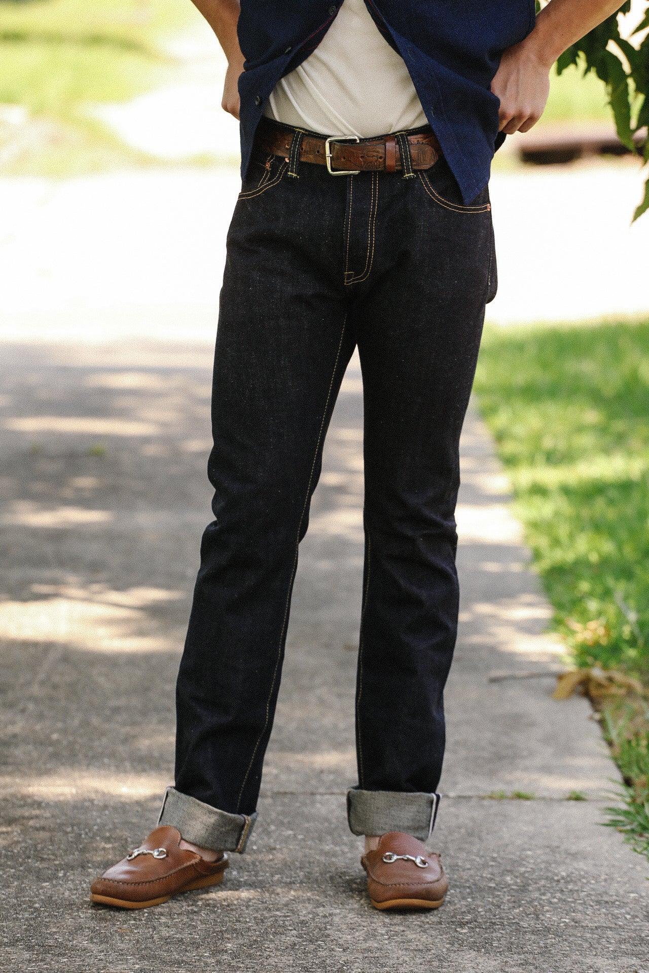 Person wearing dark denim jeans with rolled cuffs, brown leather belt, and brown loafers standing on sidewalk