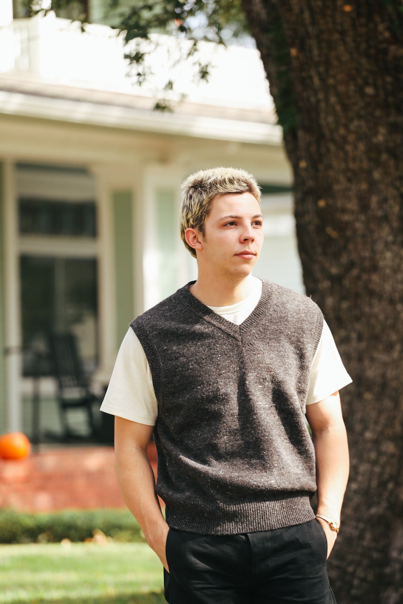 Young man wearing charcoal knit vest over white t-shirt standing outdoors by tree in front of house