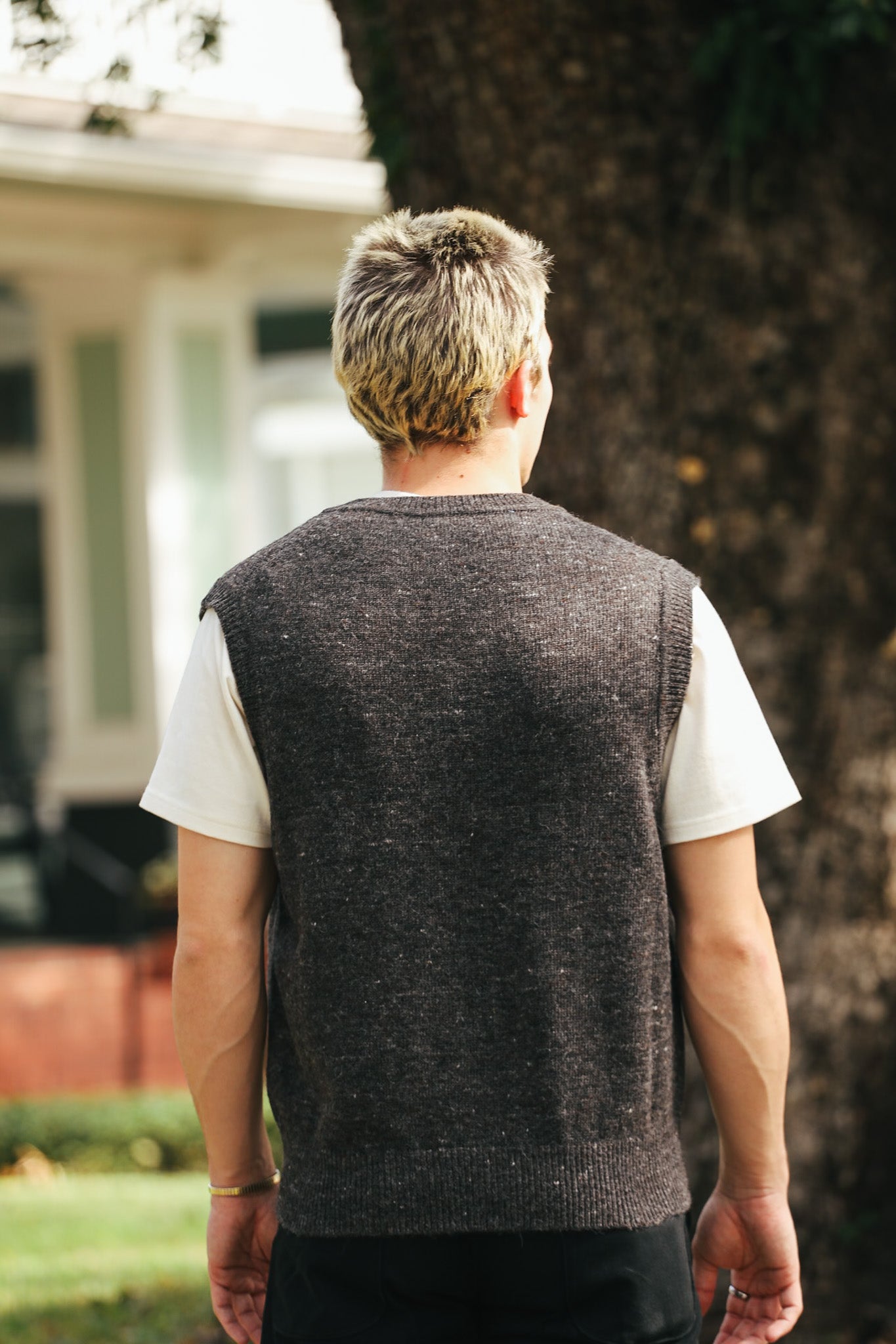 Man wearing charcoal gray knitted vest over white t-shirt standing outdoors near large tree