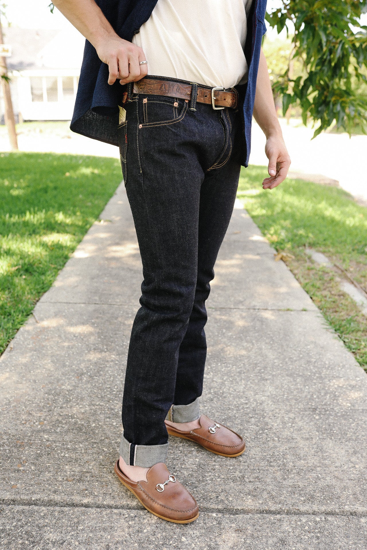 Man wearing dark denim jeans with cuffed hems, brown leather belt, and brown leather loafers outdoors