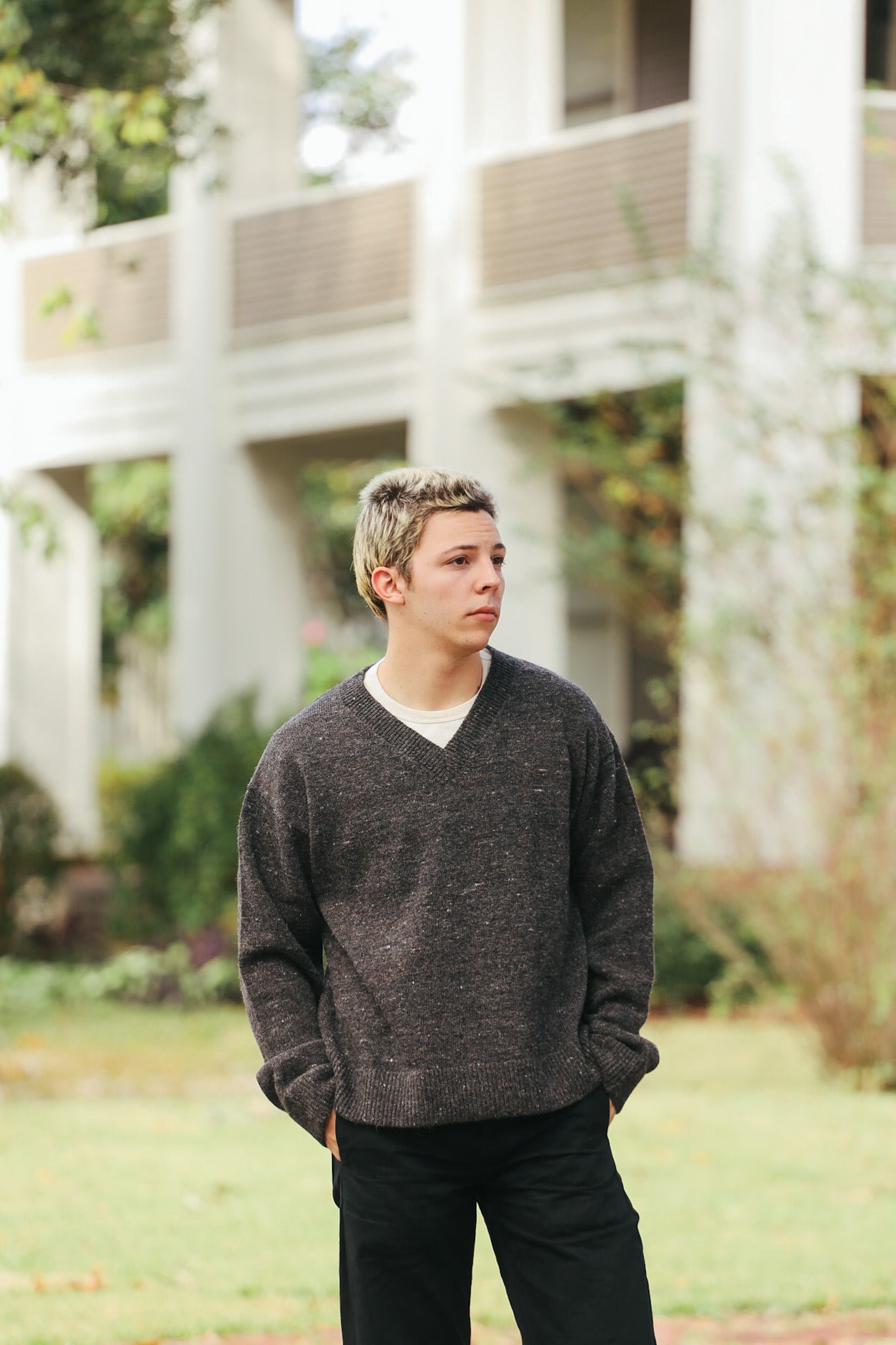 Young man wearing a dark gray V-neck sweater and black pants standing outside in a garden with a modern building in the background