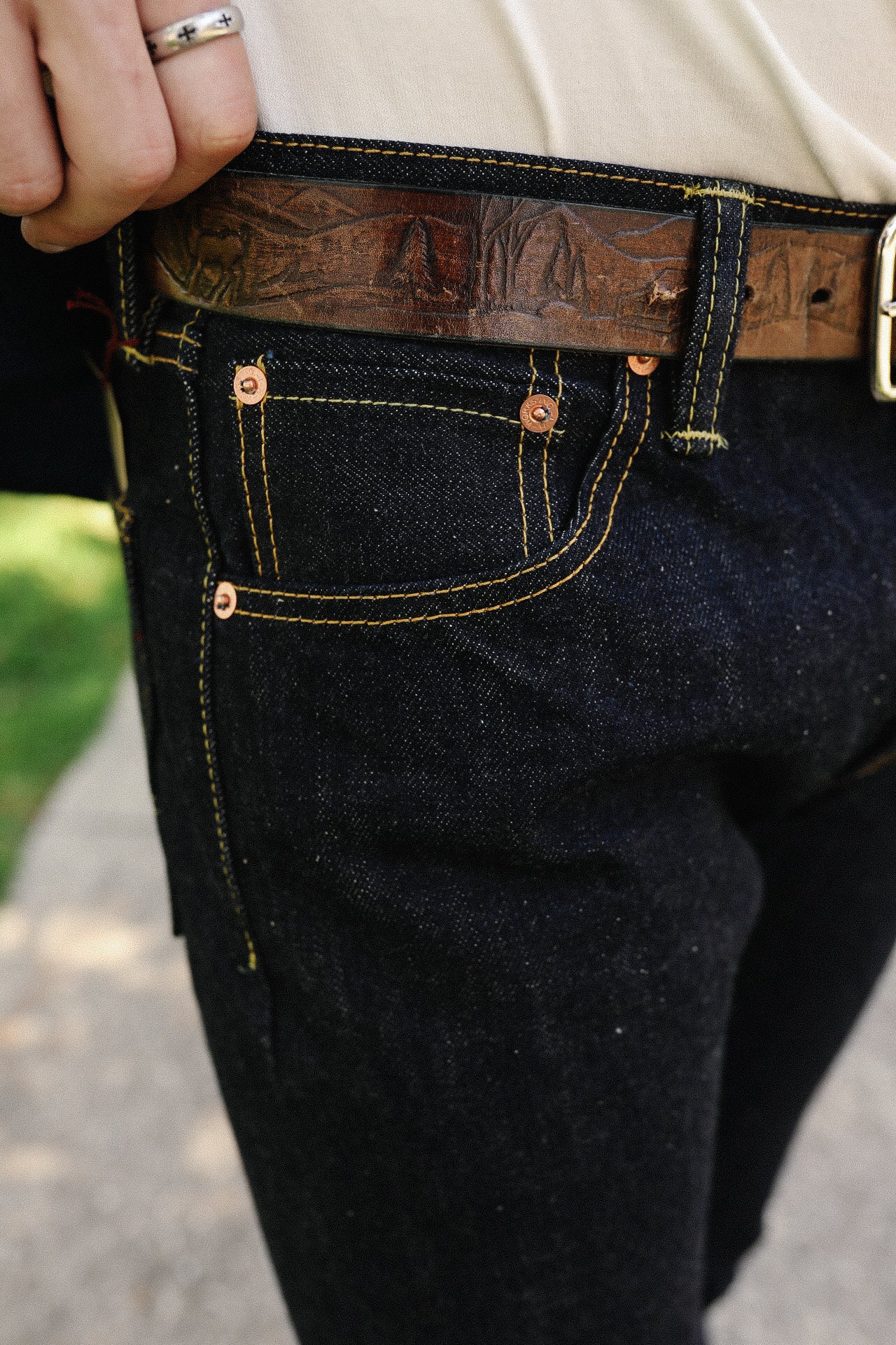 Close-up of dark denim jeans with yellow stitching and a brown embossed leather belt on a person outdoors