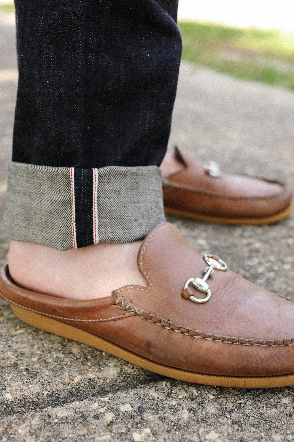 Close-up of person wearing cuffed dark denim jeans and brown leather loafers with silver horsebit detail on concrete ground