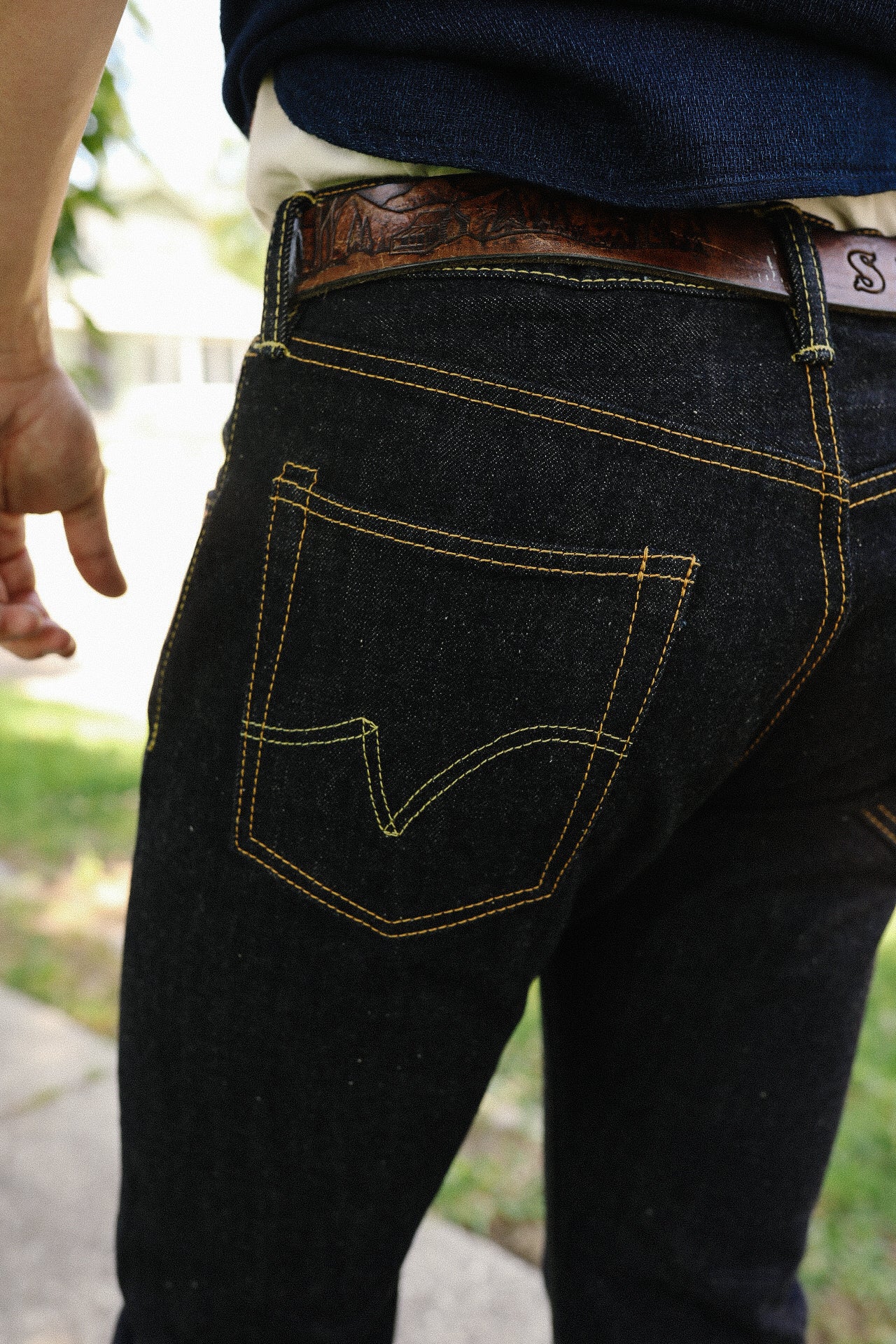 Close-up of person wearing dark denim jeans with contrast orange stitching and a brown leather belt outdoors