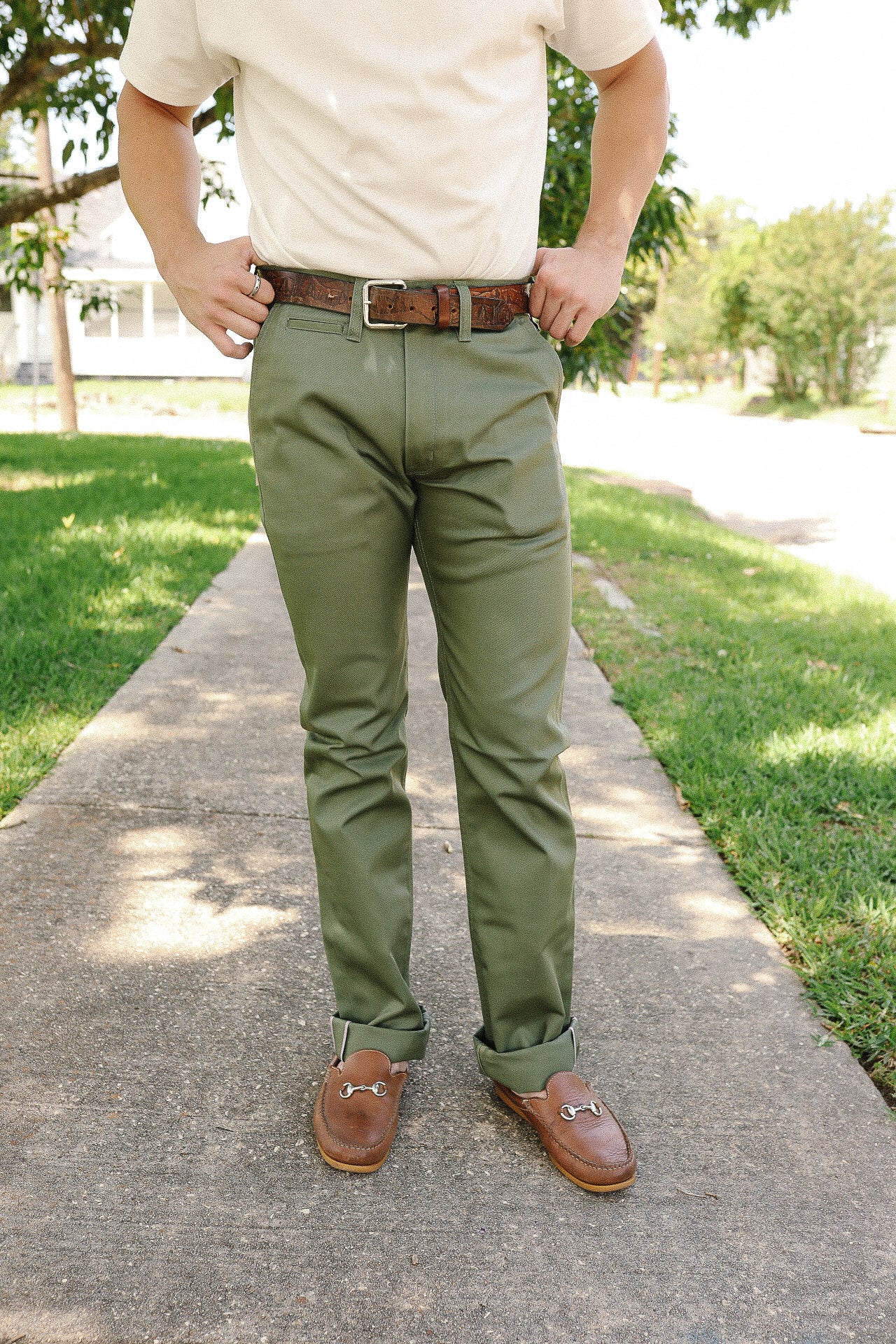 Man wearing olive green pants, brown leather belt, and brown leather loafers standing on sidewalk outdoors