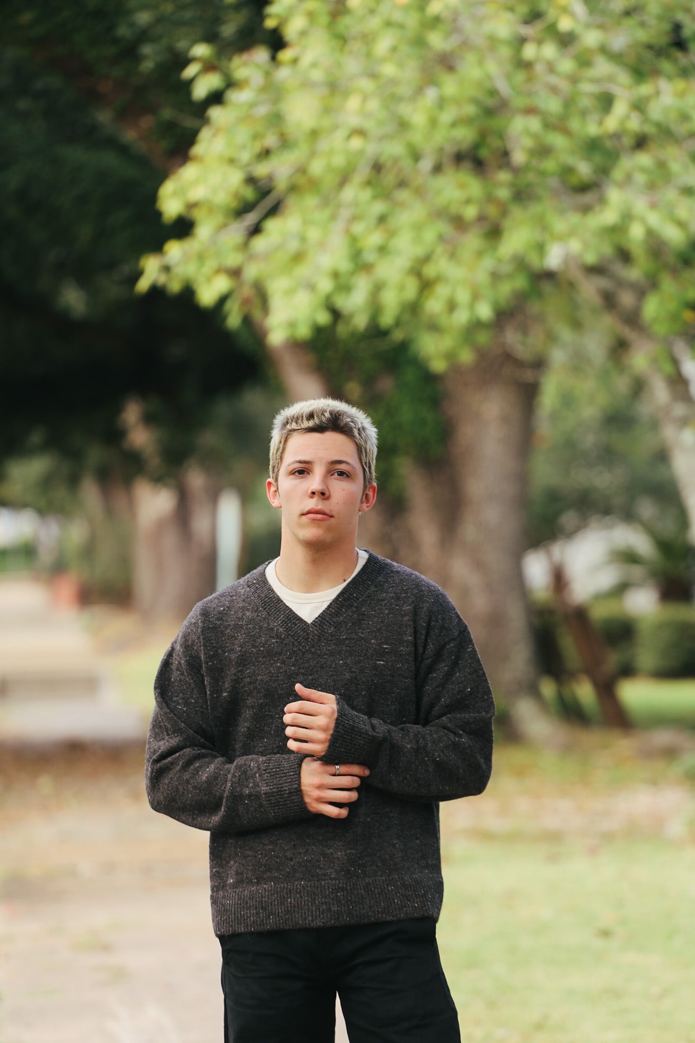 Young man wearing dark gray knit sweater standing outside with trees and walkway in background