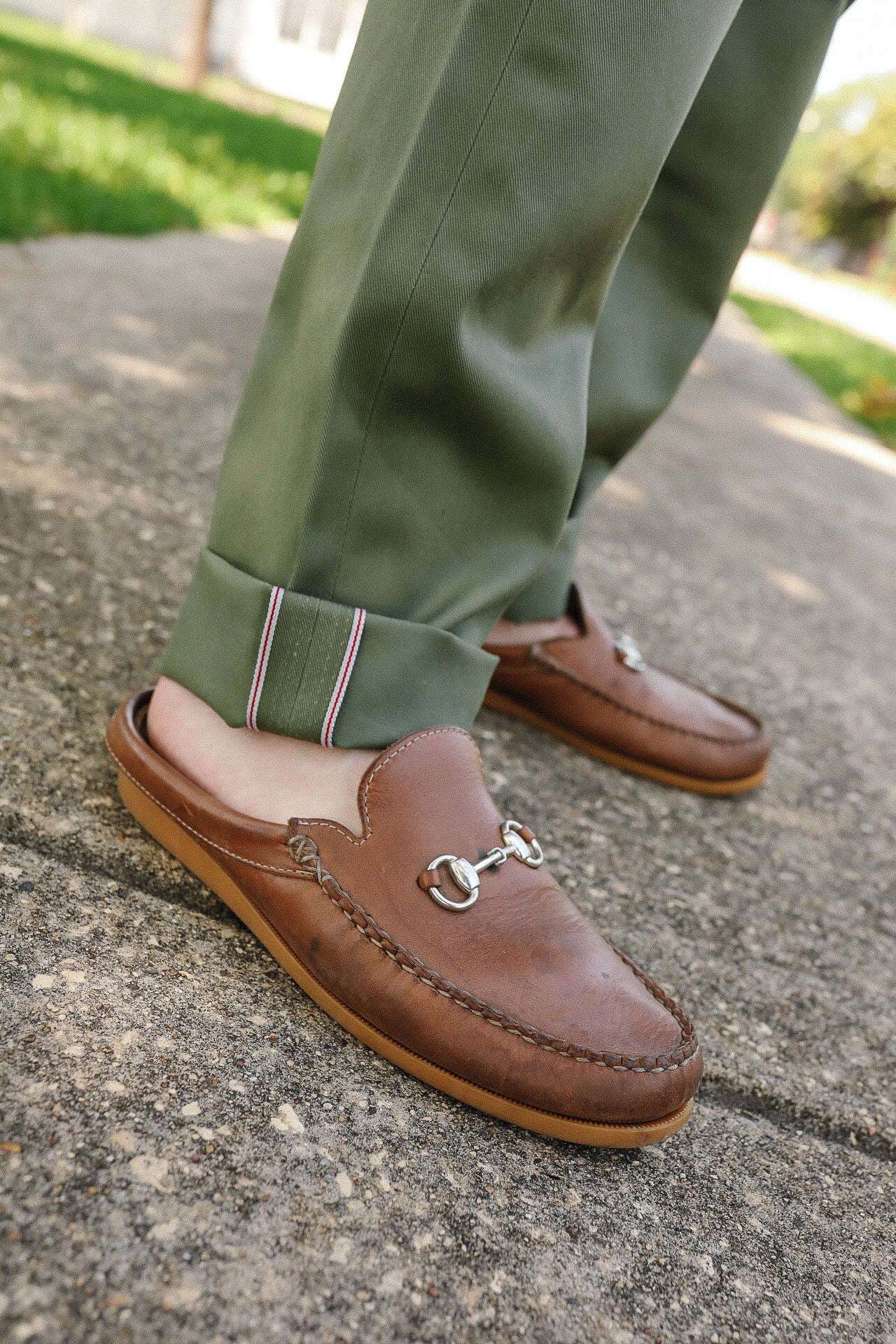 Close-up of brown leather slip-on loafers with silver bit detail worn with green cuffed pants on outdoor pavement