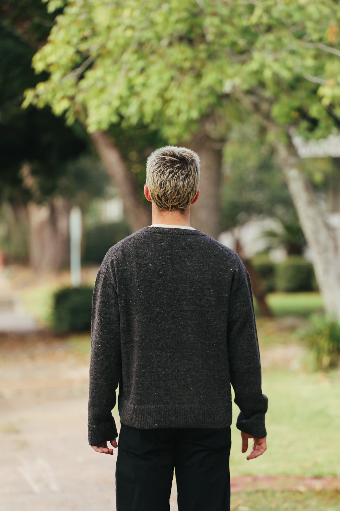Man with platinum blond hair wearing dark sweater and black pants walking outdoors under green trees