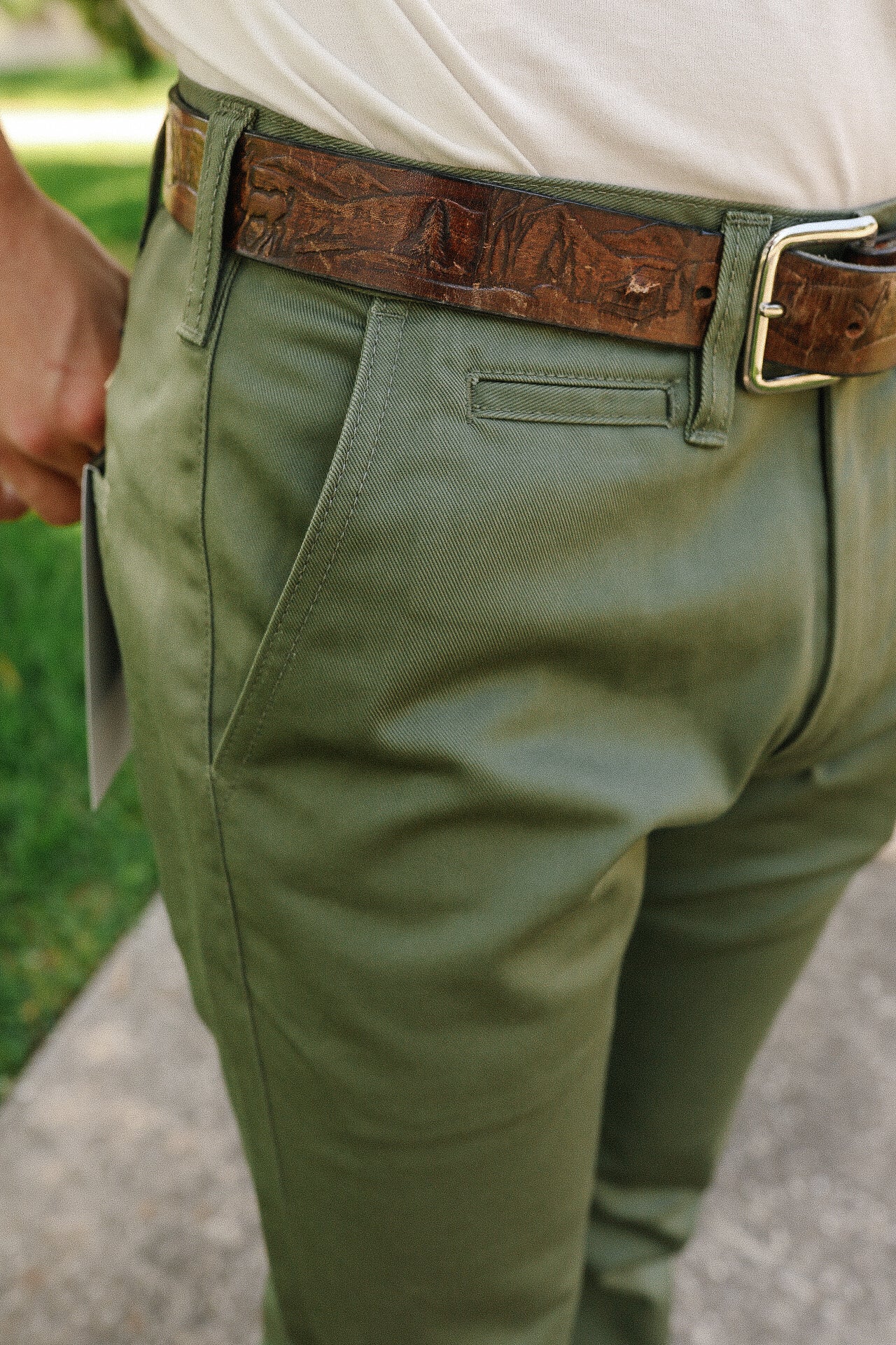 Close-up of person wearing olive green pants with a detailed brown leather belt and white shirt outdoors
