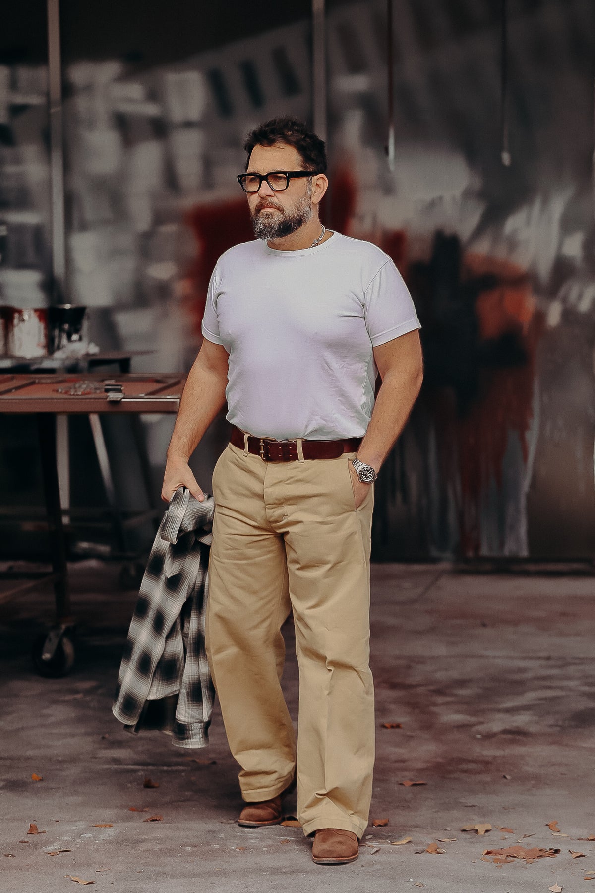 Bearded man wearing glasses, white t-shirt, beige pants, and brown boots holding plaid jacket in industrial setting
