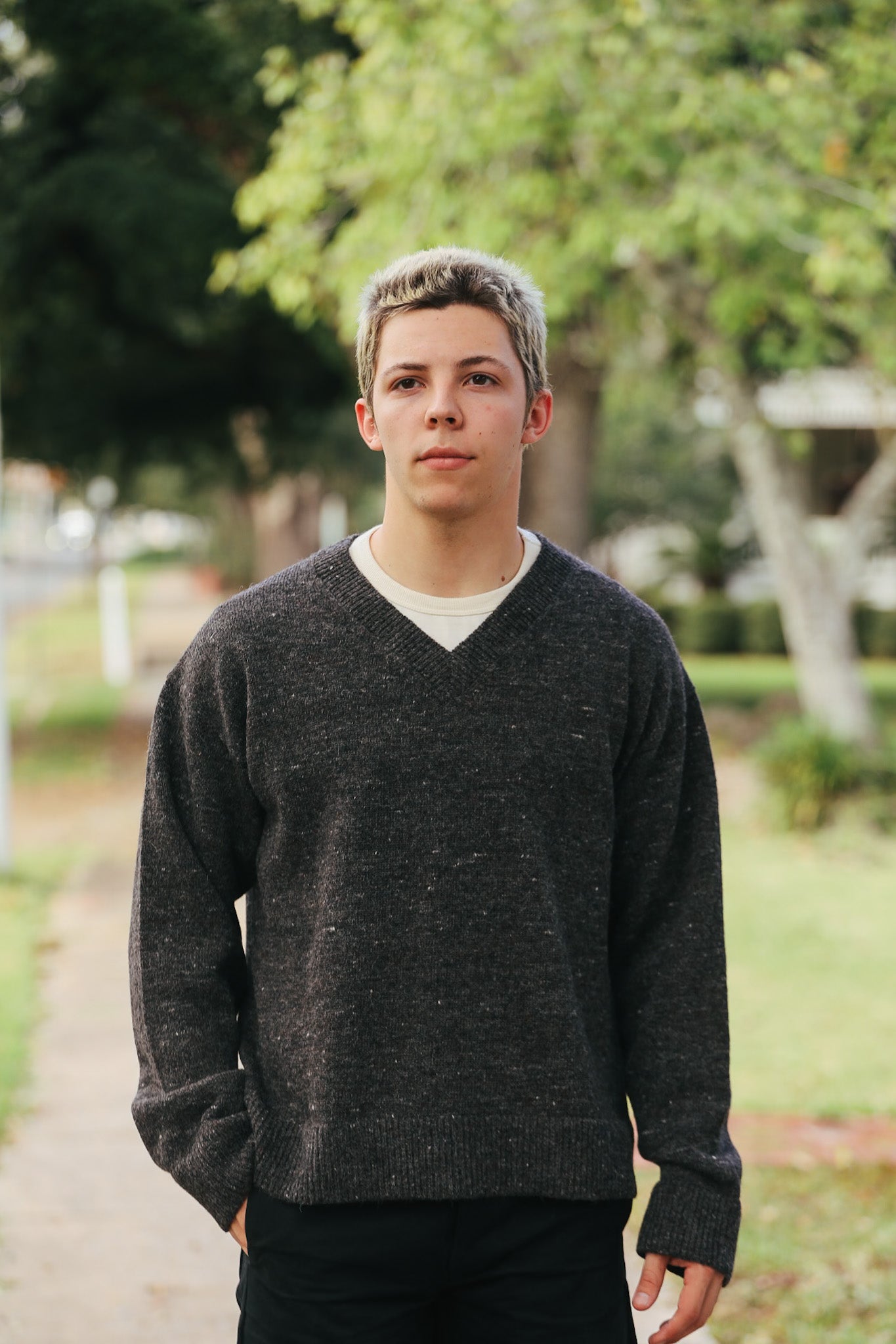 Young man wearing a dark gray V-neck sweater standing outdoors with trees and a pathway in the background
