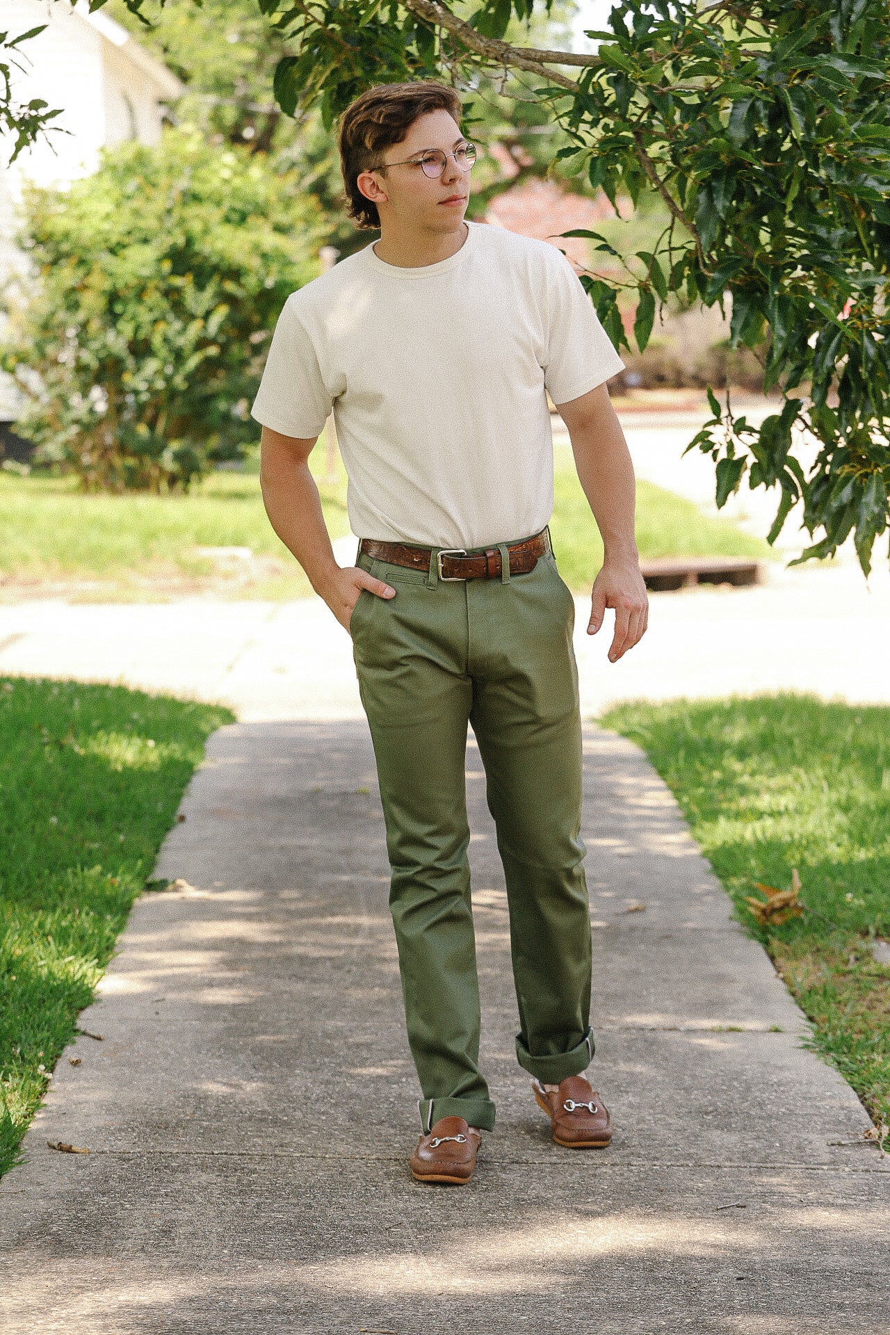Man wearing white t-shirt, olive green pants, and brown loafers walking on sidewalk in park