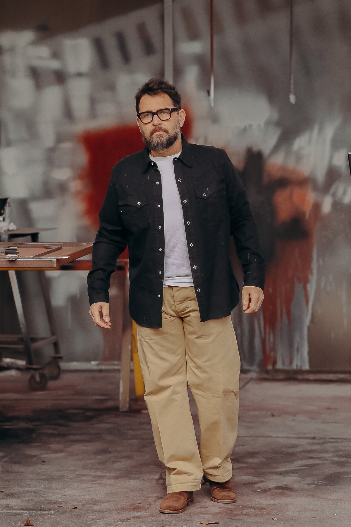 Man wearing black button-up shirt, white t-shirt, beige pants, and brown shoes in industrial workshop
